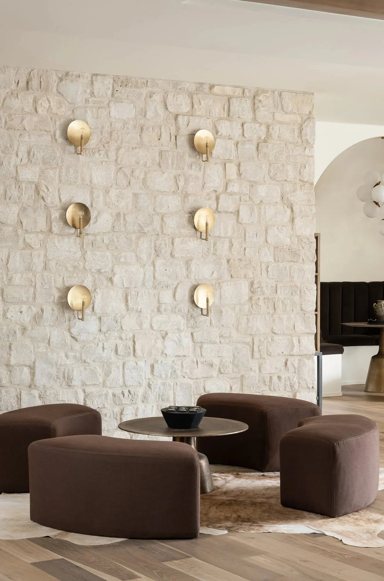 Living room with a white brick wall, wall-mounted spherical lights, a round wooden table with a black bowl, and brown upholstered chairs on a light-colored rug and wooden floor.