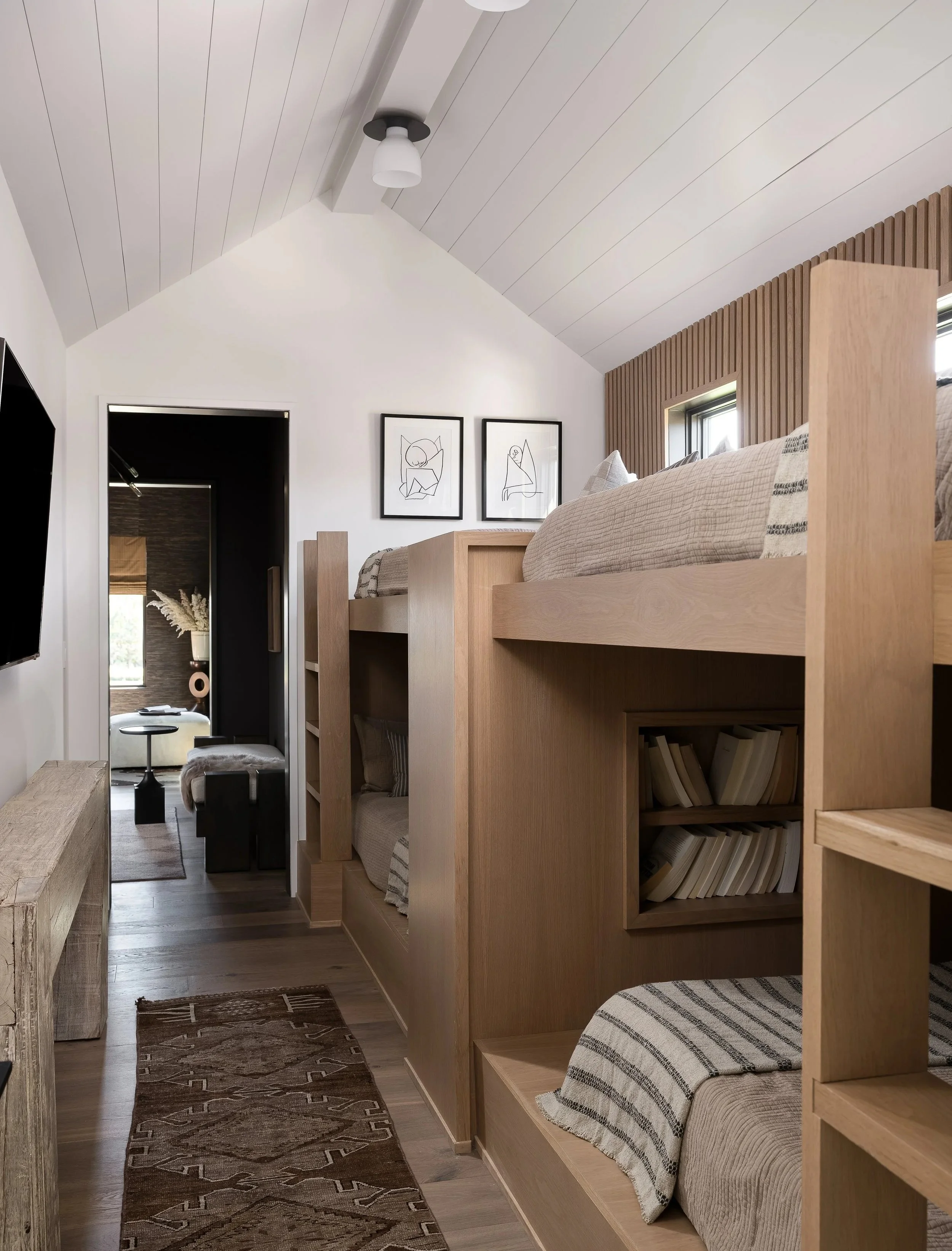 A cozy loft-style bedroom with a wooden bunk bed, beige bedding, striped pillows, a wooden bookshelf built into the bed frame, framed abstract line art on white walls, a ceiling light fixture, and windows letting in natural light.