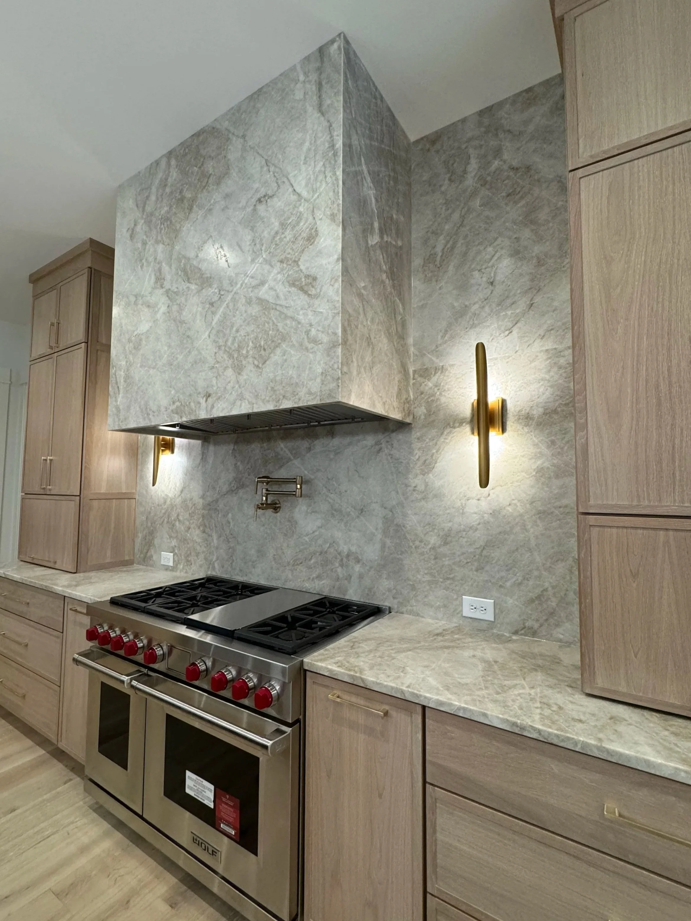 Modern kitchen with a built-in stove with red control knobs, beige countertops, and wooden cabinets. There is a gray marble backsplash and an exhaust hood. Two gold wall-mounted lights are on the wall.