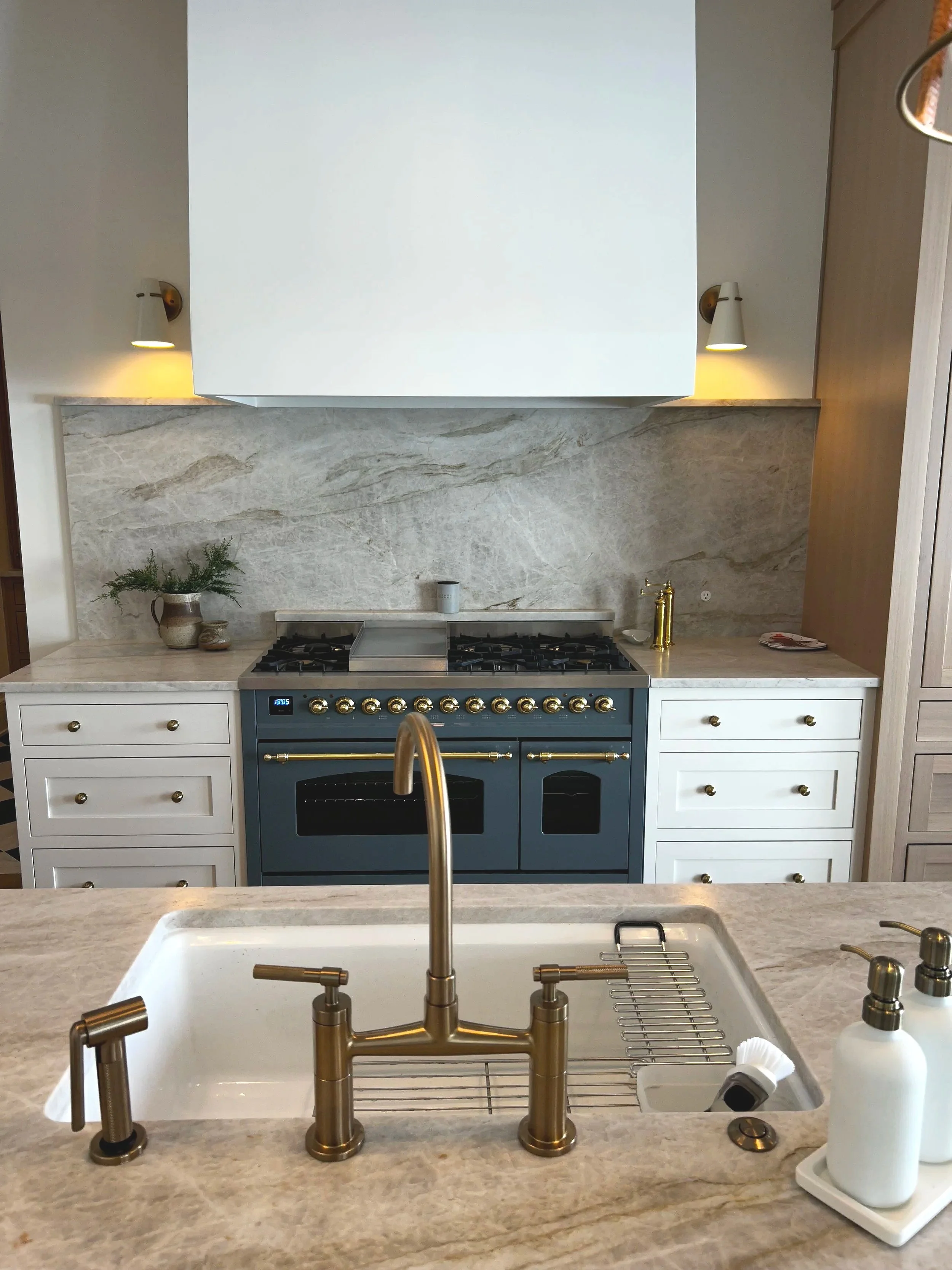 Kitchen view from the countertop looking towards the stove and backsplash with a sink in the foreground. The backsplash is made of light-colored stone, and there are small plants and bottles on the counter.