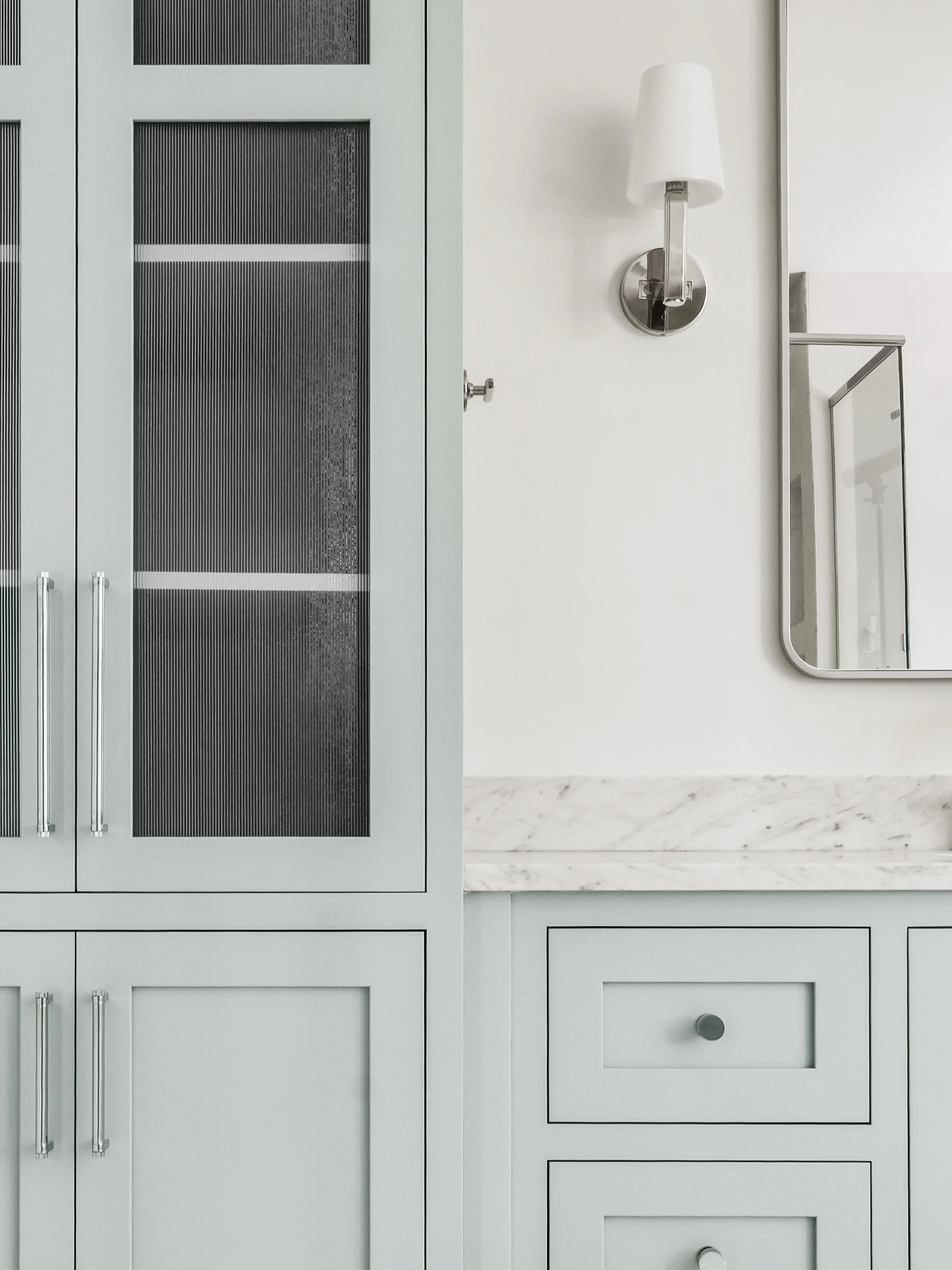 Kitchen cabinets with glass-paneled doors, marble countertop, mirror, and wall-mounted light fixture.