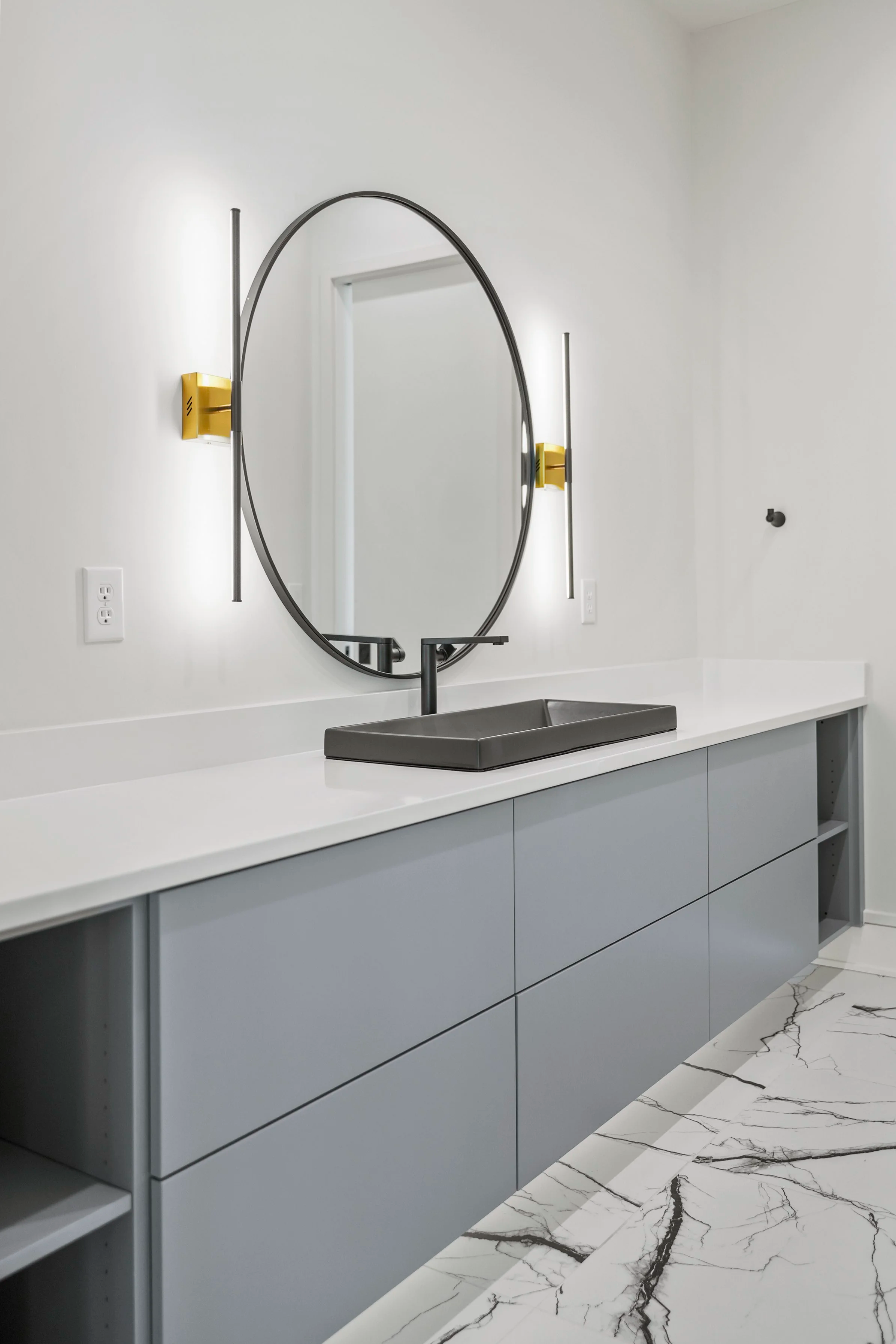 Modern bathroom sink area with a large round mirror, black sink basin, white countertop, and light gray cabinets below. Two vertical light fixtures with yellow accents are mounted on the wall, and the floor features white marble tiles with black vein