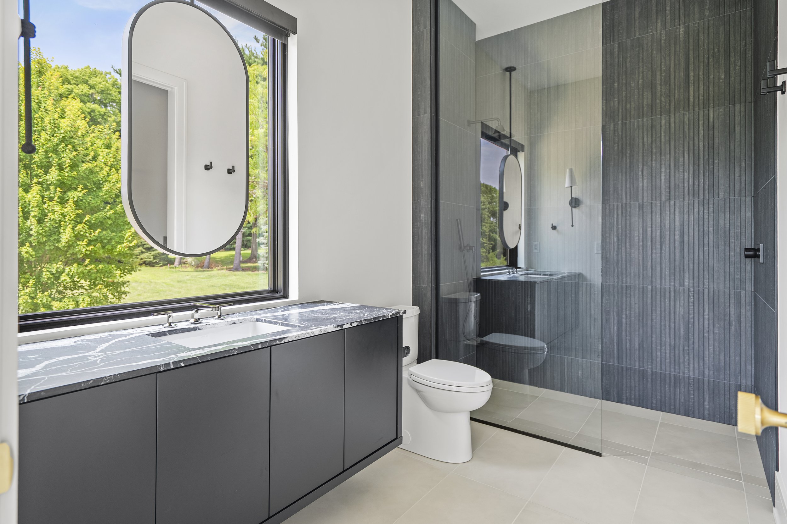 Modern bathroom with gray vanity, marble countertop, large window overlooking greenery, gray tiled shower with glass door, toilet, and wall-mounted fixtures.