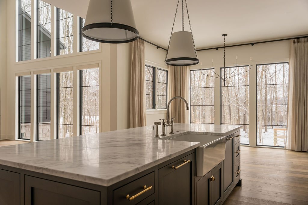 Modern kitchen with marble island, black cabinets, gold hardware, large windows, and pendant lighting.