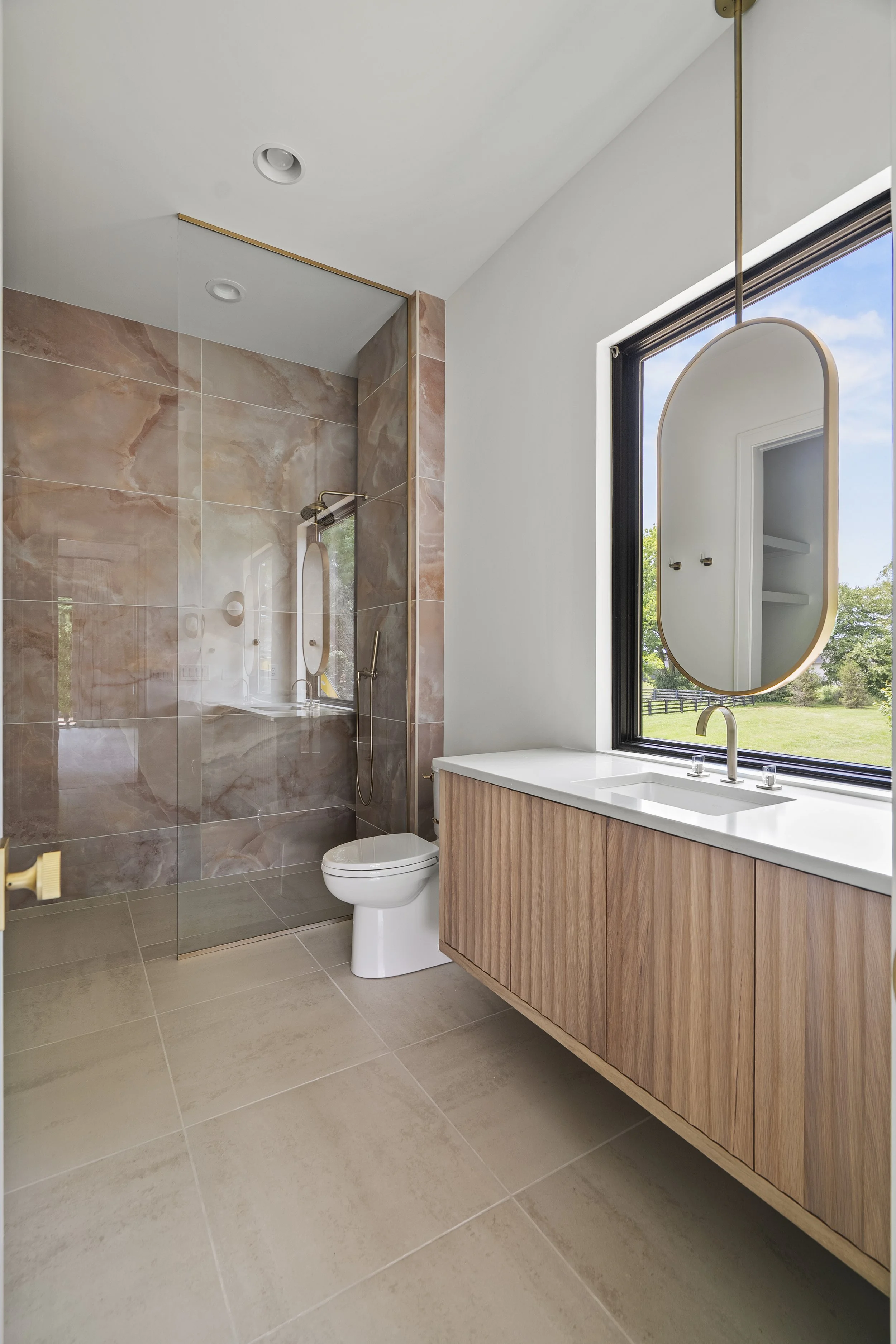Modern bathroom with a glass-enclosed shower area with marble tiles, a white toilet, a wooden cabinet with a white countertop and integrated sink, a large window with a view of green landscape, and a decorative oval mirror hanging beside the window.