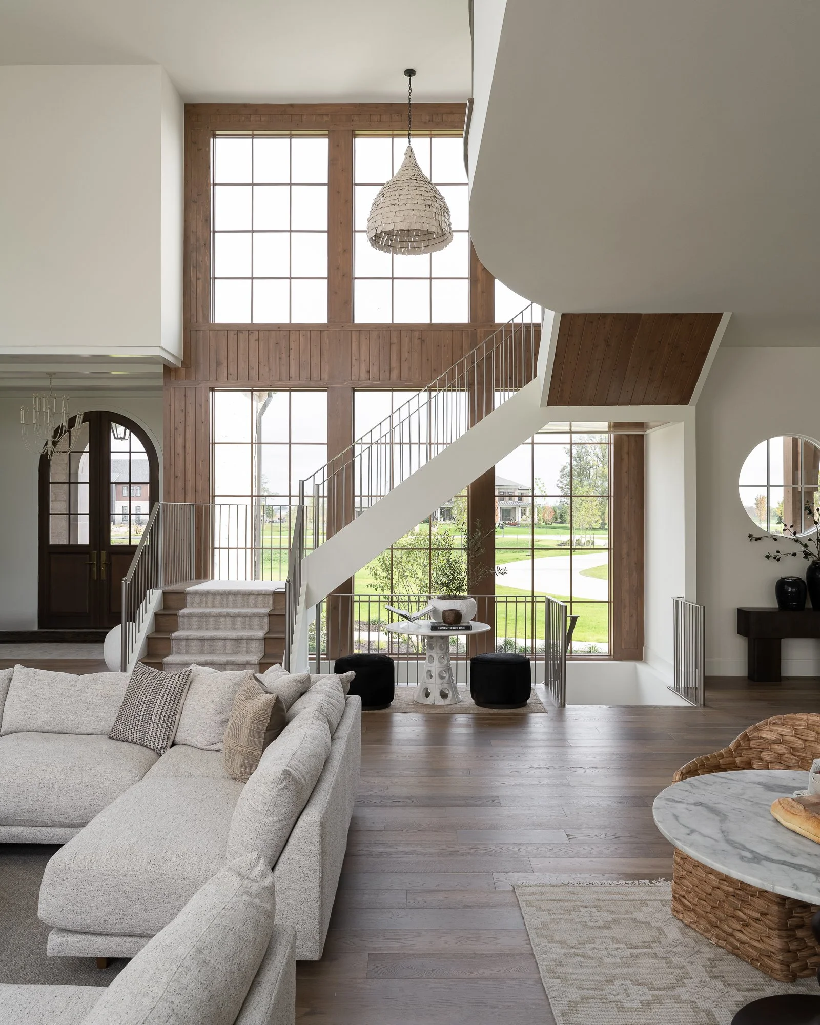 Living room with large window, staircase, beige sofa, round marble-topped table, woven baskets, and wood accents.