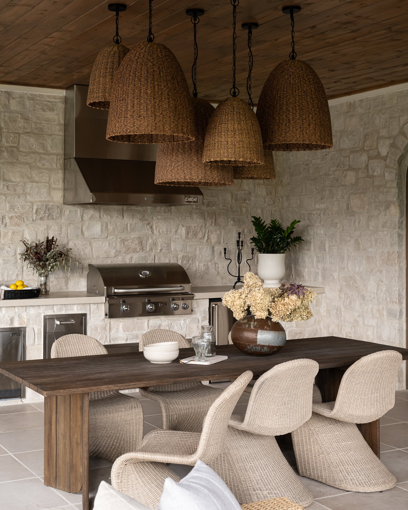 Interior of a dining area with a rustic wooden table, woven chairs, and a large vase of cream flowers, adjacent to a kitchen with a stone wall and a stainless steel range hood.