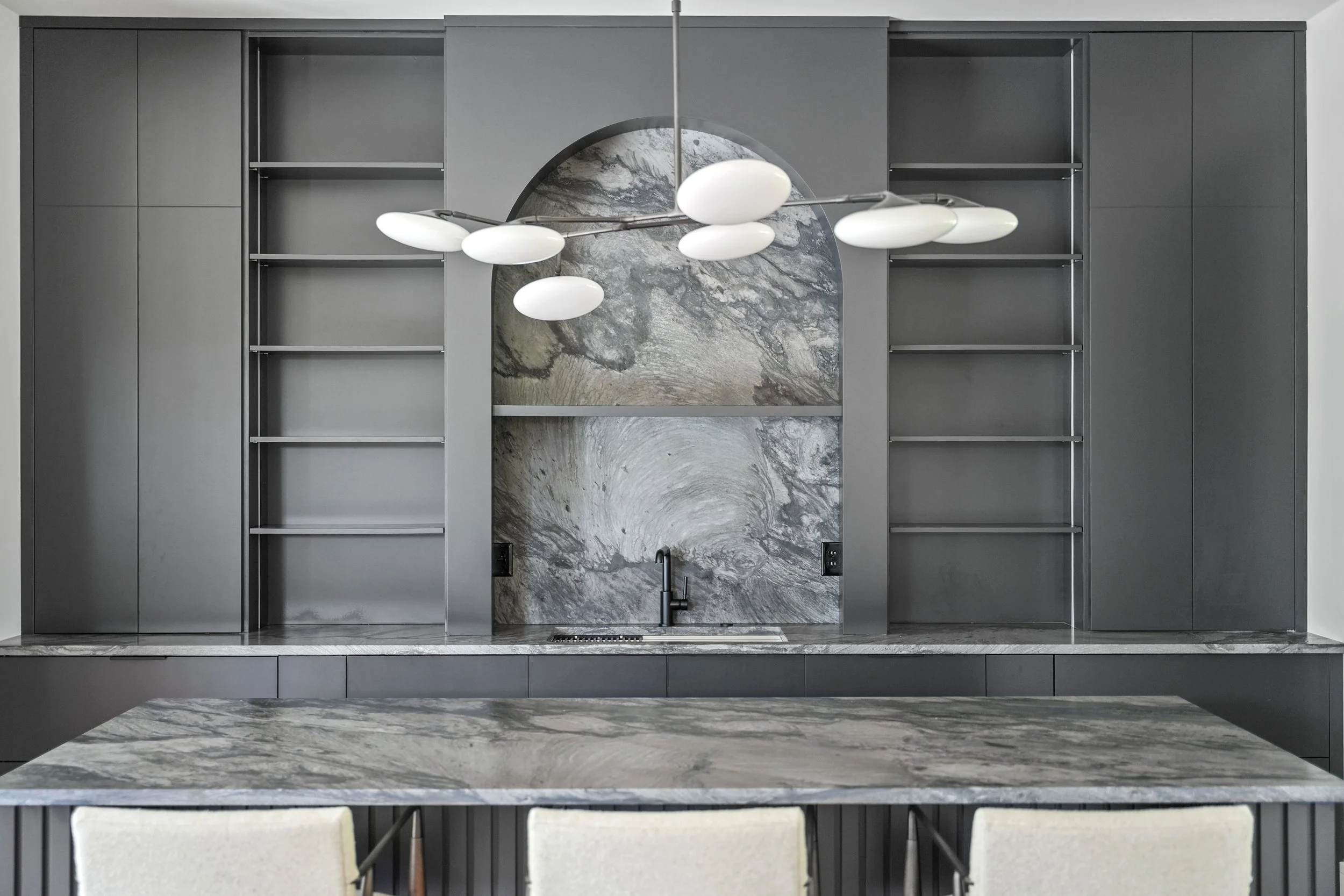 Interior view of a modern kitchen with dark gray cabinets, marble backsplash, a sink with a black faucet, and a sleek hanging light fixture.
