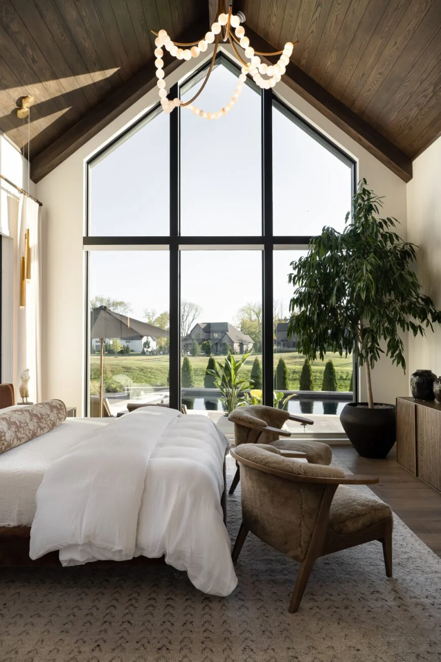 Modern bedroom with large floor-to-ceiling window showcasing a green outdoor landscape. The room features a white bed with a wooden headboard, two beige armchairs, a potted plant, and a chandelier hanging from the wooden ceiling.