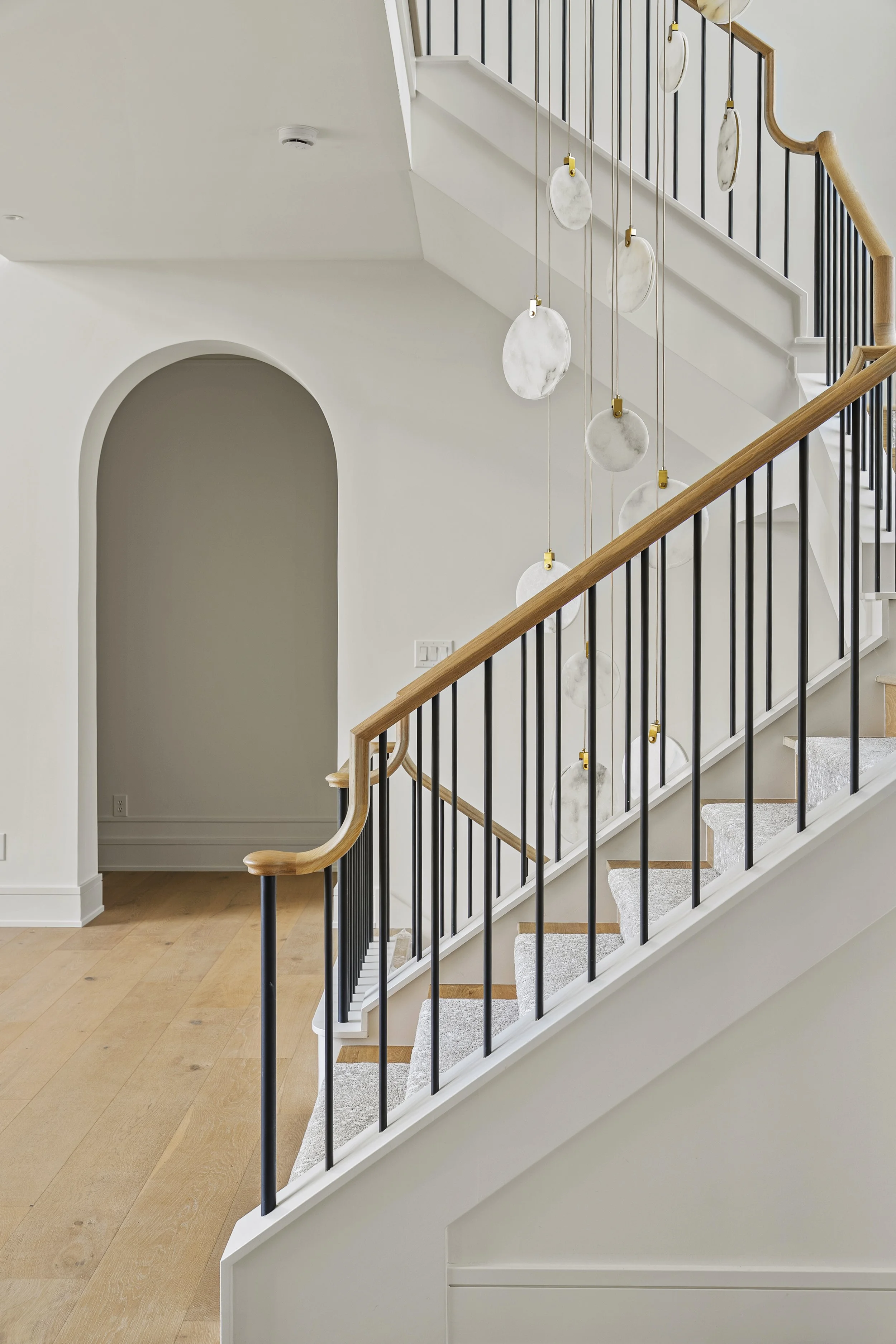 Interior of a modern staircase with white walls, wooden handrails, black metal balusters, and a hanging light fixture with evenly spaced circular white shades.