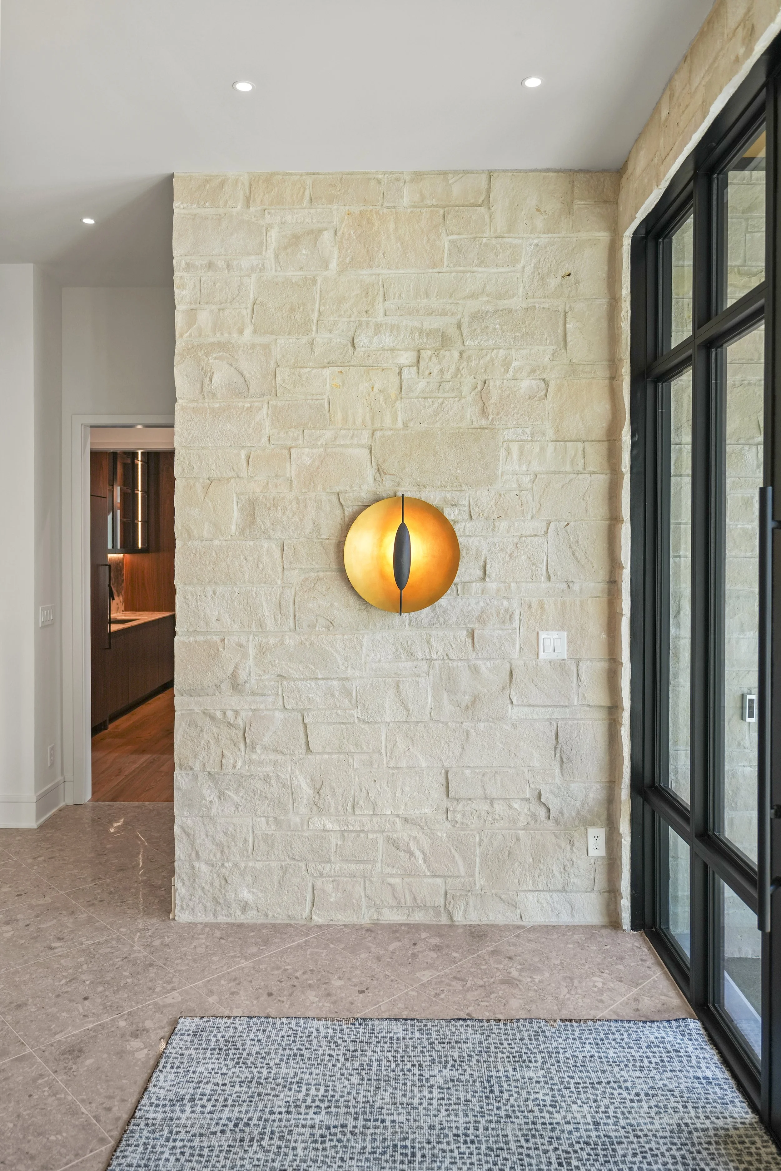 Interior of a modern home with beige stone wall, wall-mounted light fixture, large windows with black frames, and a doorway showing a kitchen area.