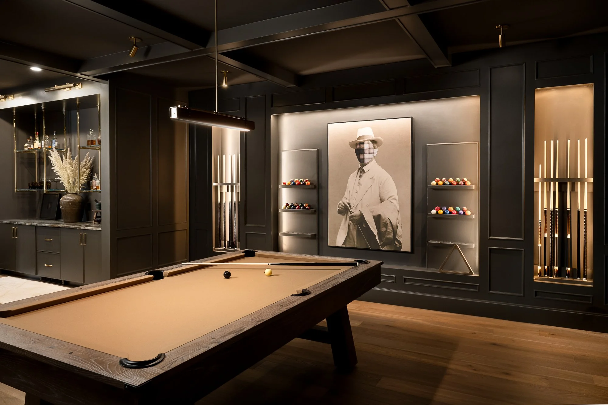 Interior of a modern game or recreational room with a pool table, wall art of a person with a checkered face mask and a hat, and shelves with billiard balls, cue sticks, and bar supplies, all in a dark-toned decor with subtle lighting.