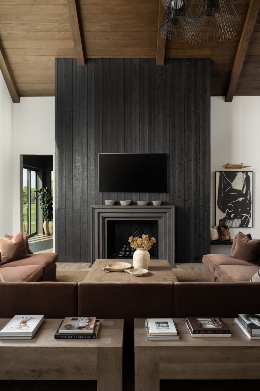 Living room with a wooden ceiling, black accent wall with a mounted TV, a fireplace with decorative objects, beige sofas, a wooden coffee table with a flower vase, and books on a wooden side table.