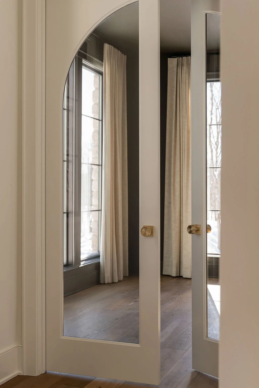 A pair of white closet doors with glass panels and gold handles, slightly open, reflecting a room with large windows, beige curtains, and wooden flooring.