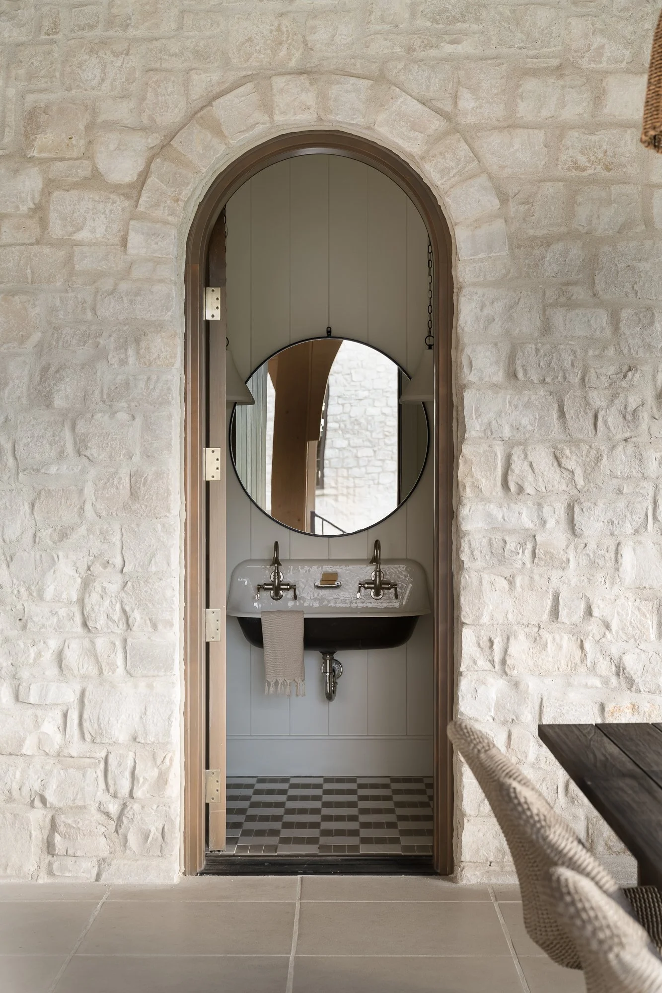 A doorway with a brick arch opening into a small bathroom with a round mirror, a sink, and a towel hanging on the side. The bathroom interior features a checkered tile floor.