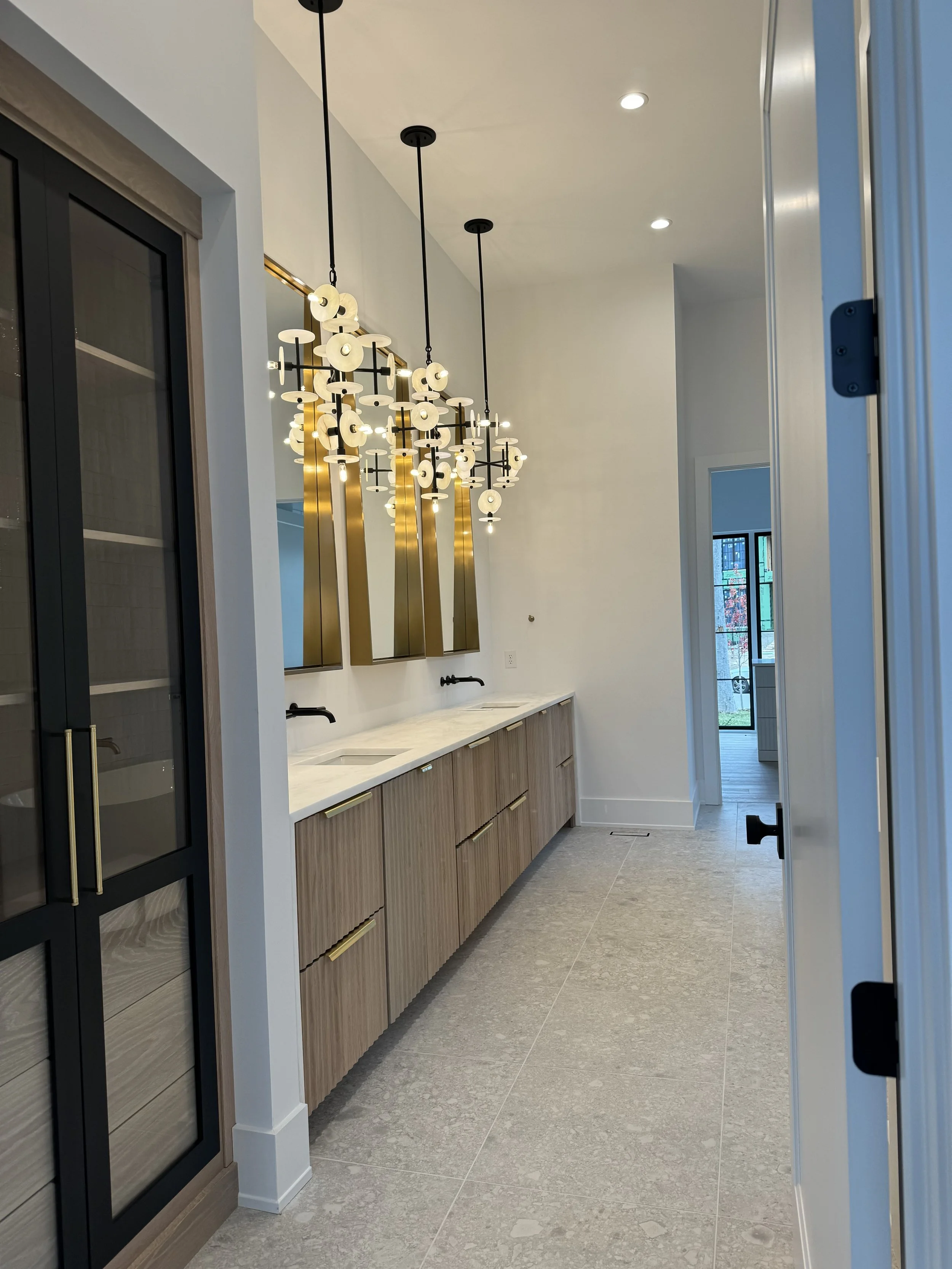 Modern bathroom with stone tile flooring, a long wooden vanity with a marble countertop, black faucets, and a large contemporary chandelier with multiple lights and disc-shaped shades. There is a window looking outside and a mirrored wall behind the 