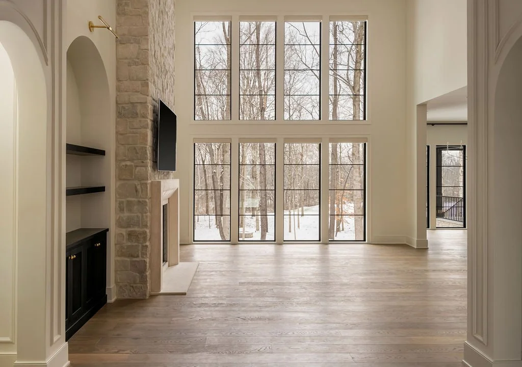 Empty living room with large windows overlooking a snowy outdoor scene, featuring a stone fireplace, a wall-mounted TV, built-in shelves, and light hardwood floors.