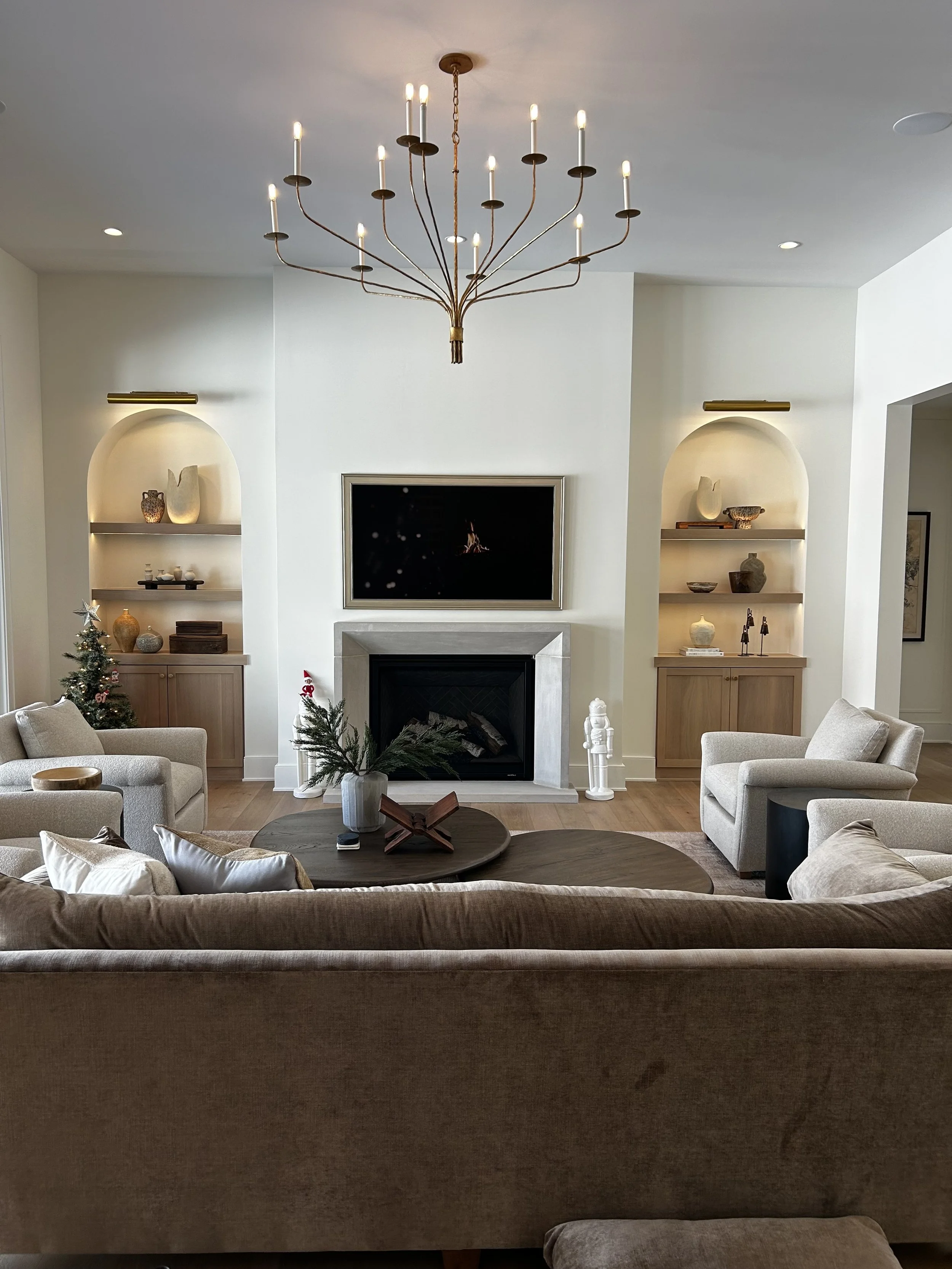 Living room with beige sofa, two armchairs, coffee table, fireplace, wall-mounted TV, built-in shelves with decorative items, small Christmas tree, and modern chandelier overhead.