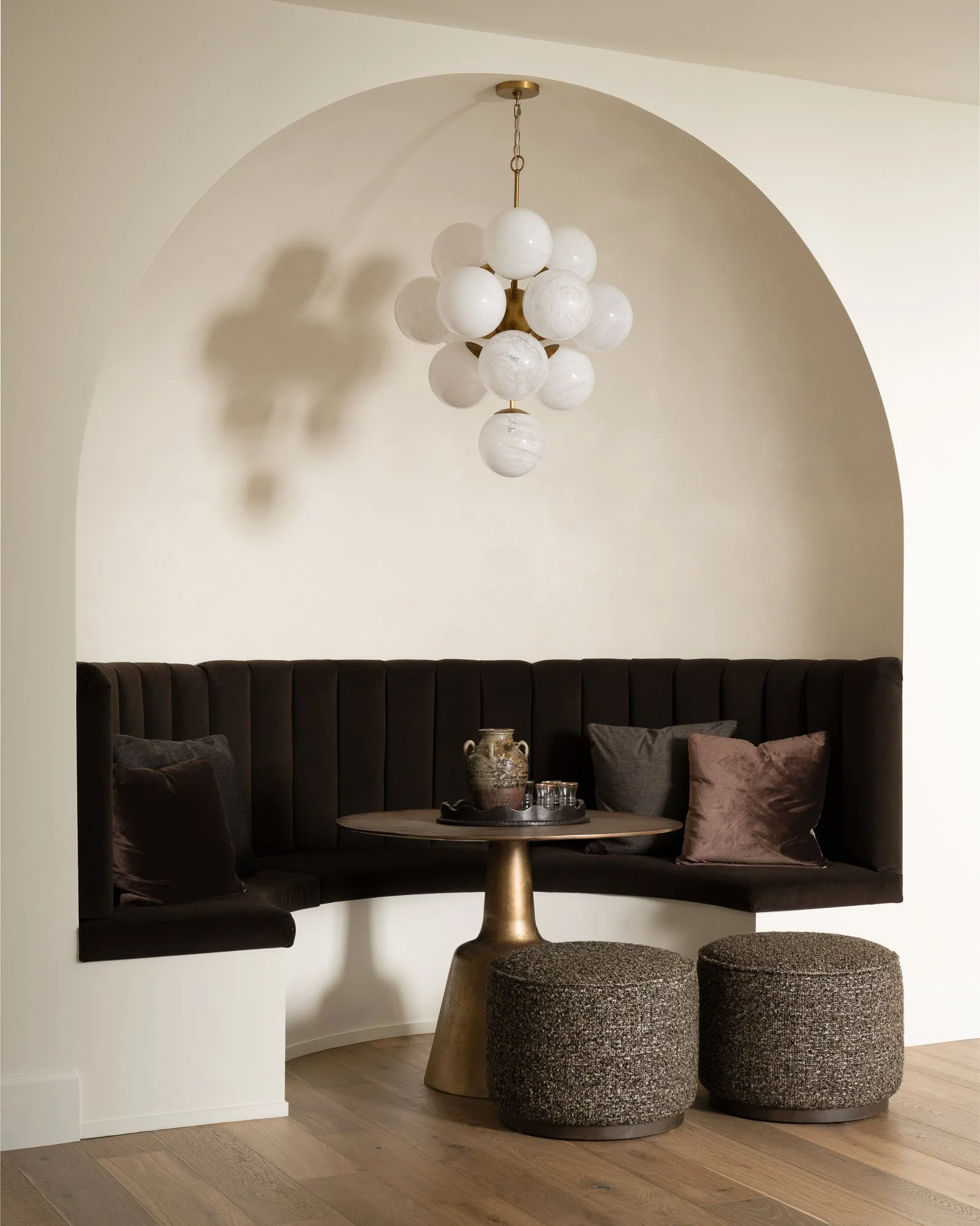 A cozy dining nook with a black, upholstered curved banquette seating area, mustard-colored table, and textured gray ottomans. A decorative metallic pitcher and tray with glasses sit on the table. An ornate chandelier with multiple spherical white gl