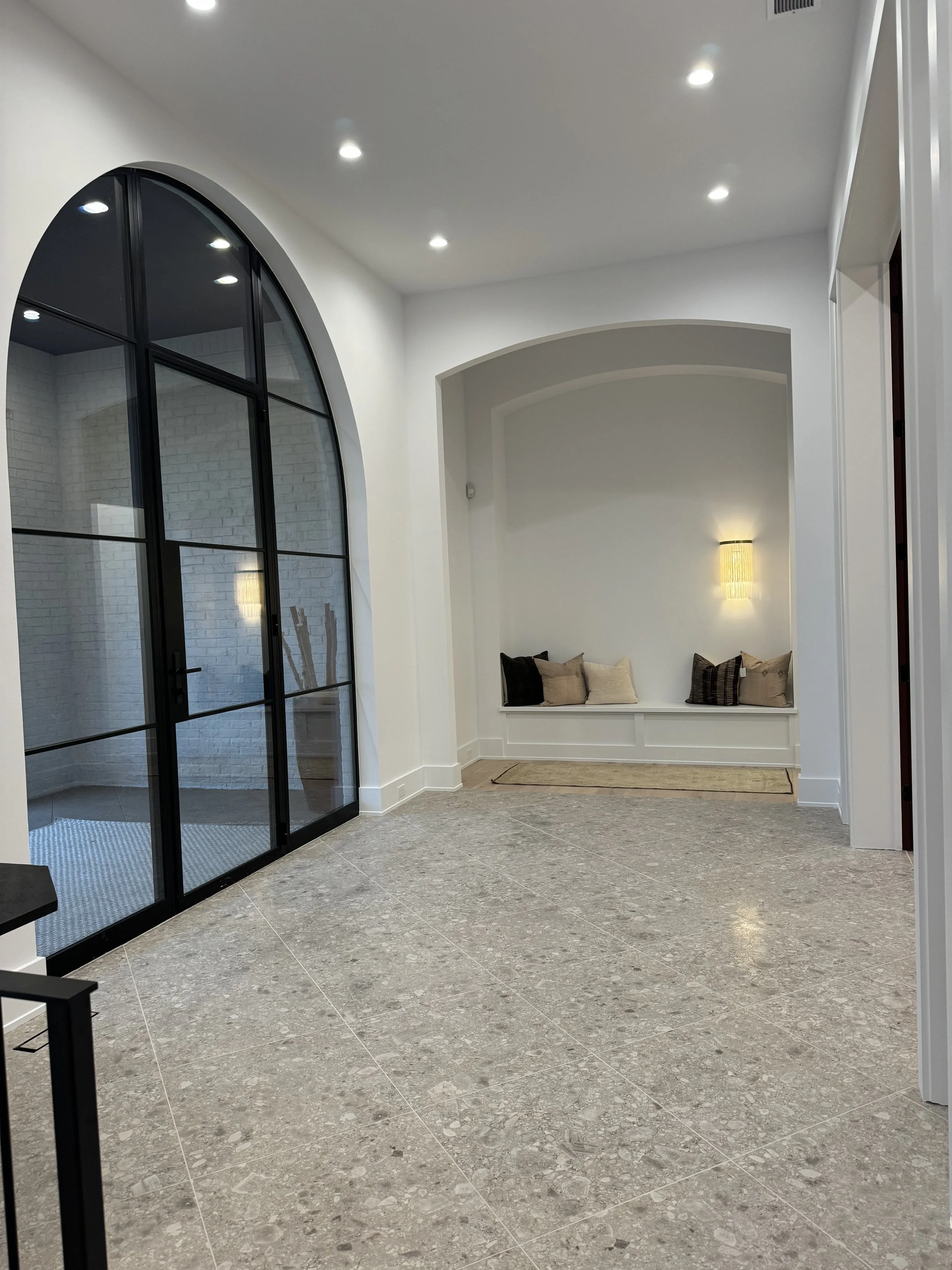 Interior of a modern room with graystone tile flooring, white walls, a large arched window with black framing, and a built-in window seat with pillows. Recessed ceiling lights and a wall-mounted lamp illuminate the space.