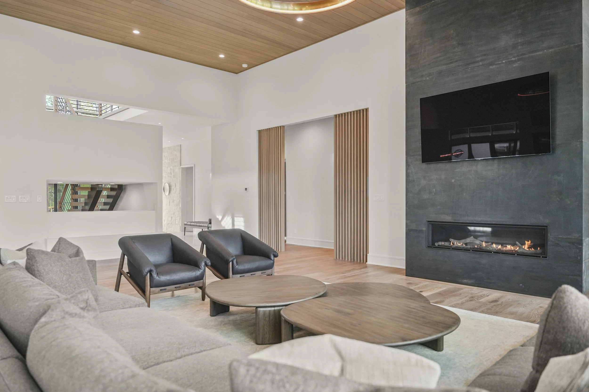 Modern living room featuring a fireplace with a black TV mounted above, black armchairs, a gray sofa with pillows, and wooden coffee tables, with wooden ceiling panels and light-colored hardwood flooring.