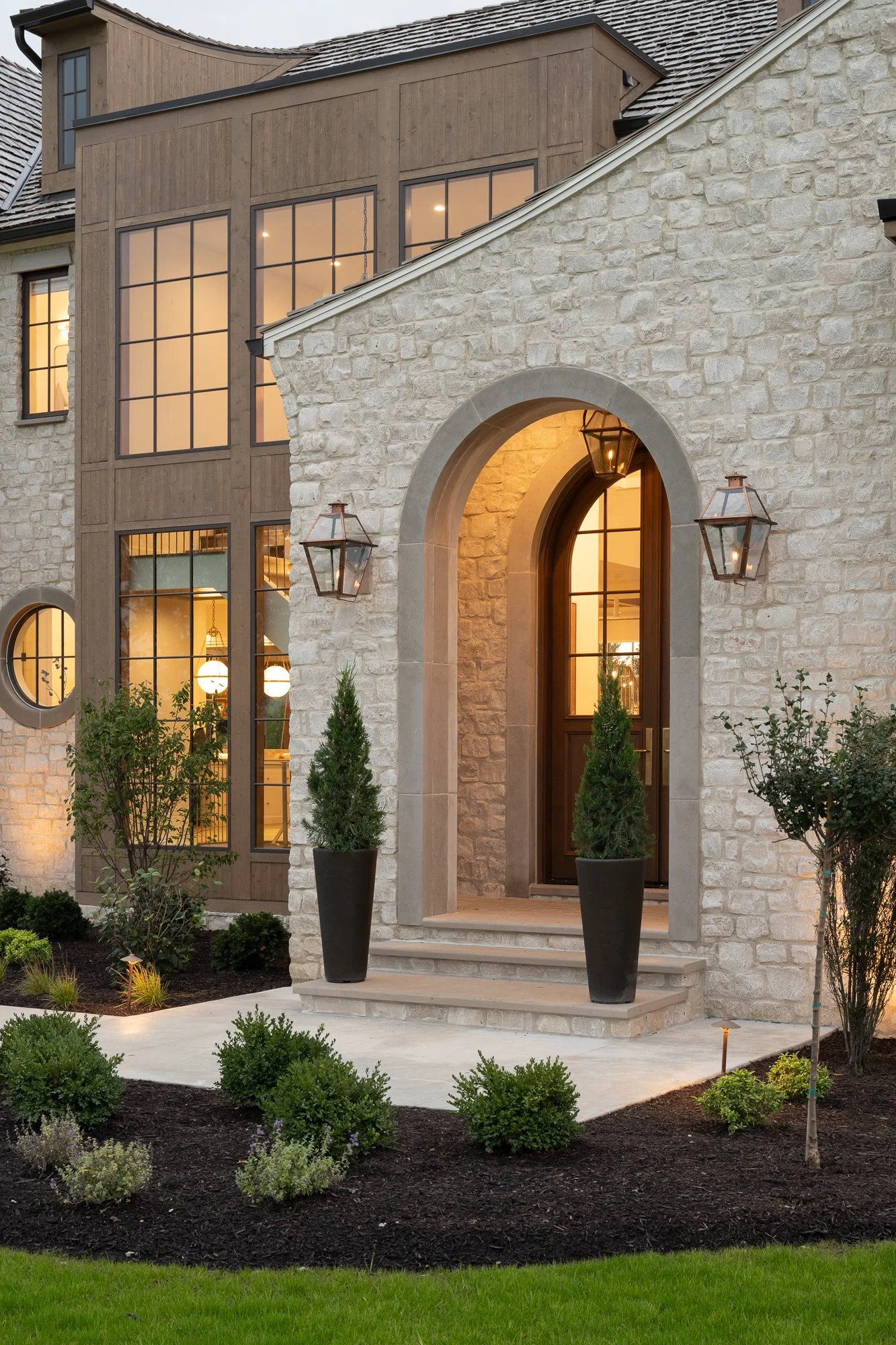 Front view of a modern house with stone and wood exterior, large windows, potted plants at the entrance, and landscaped garden with shrubs and grass.