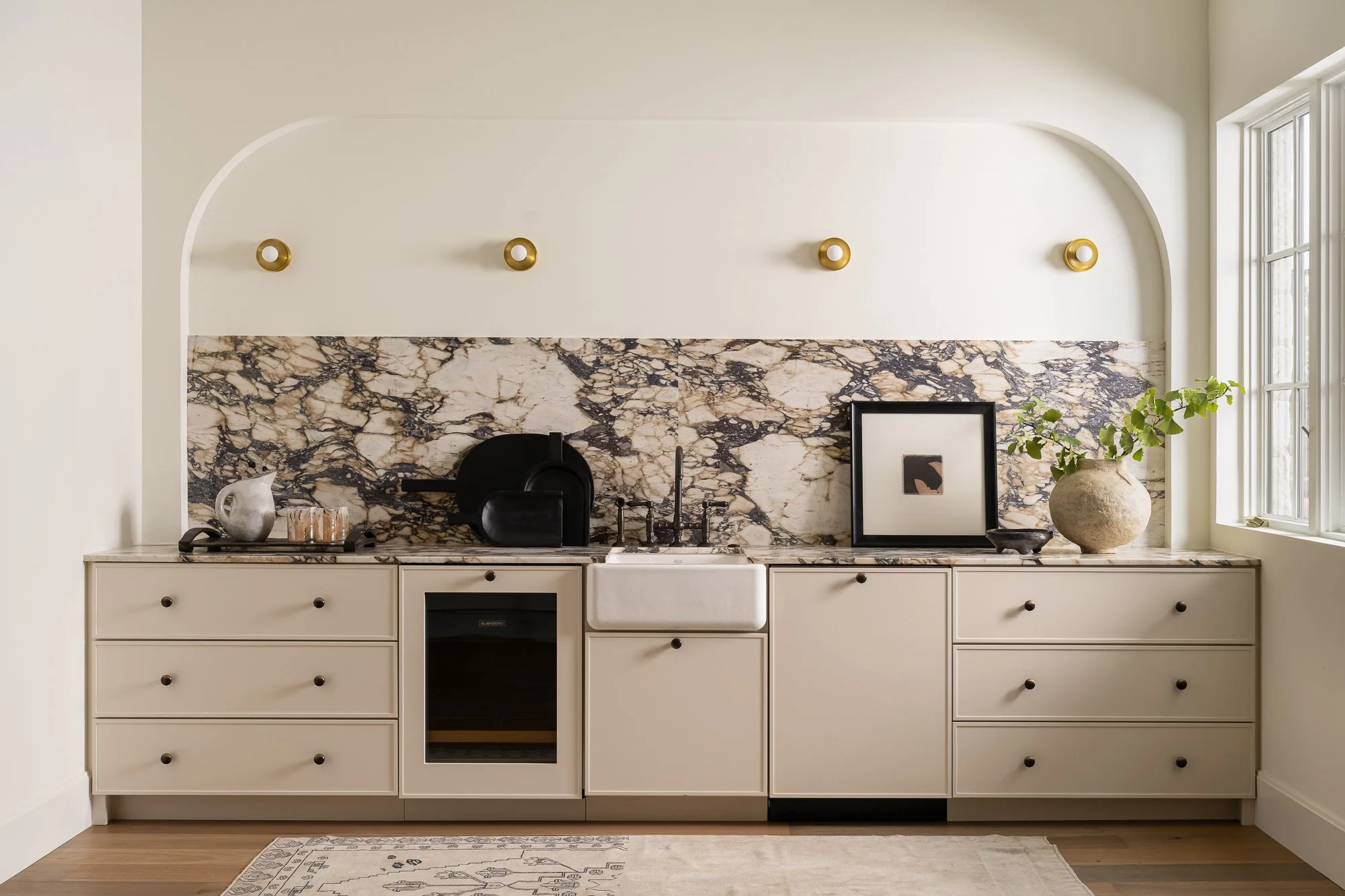 Kitchen with cream-colored cabinets, marble backsplash, window on the right, gold wall hooks, decorative items including a silver pitcher, glassware, framed artwork, and a large beige vase with green plant, situated on a marble countertop.