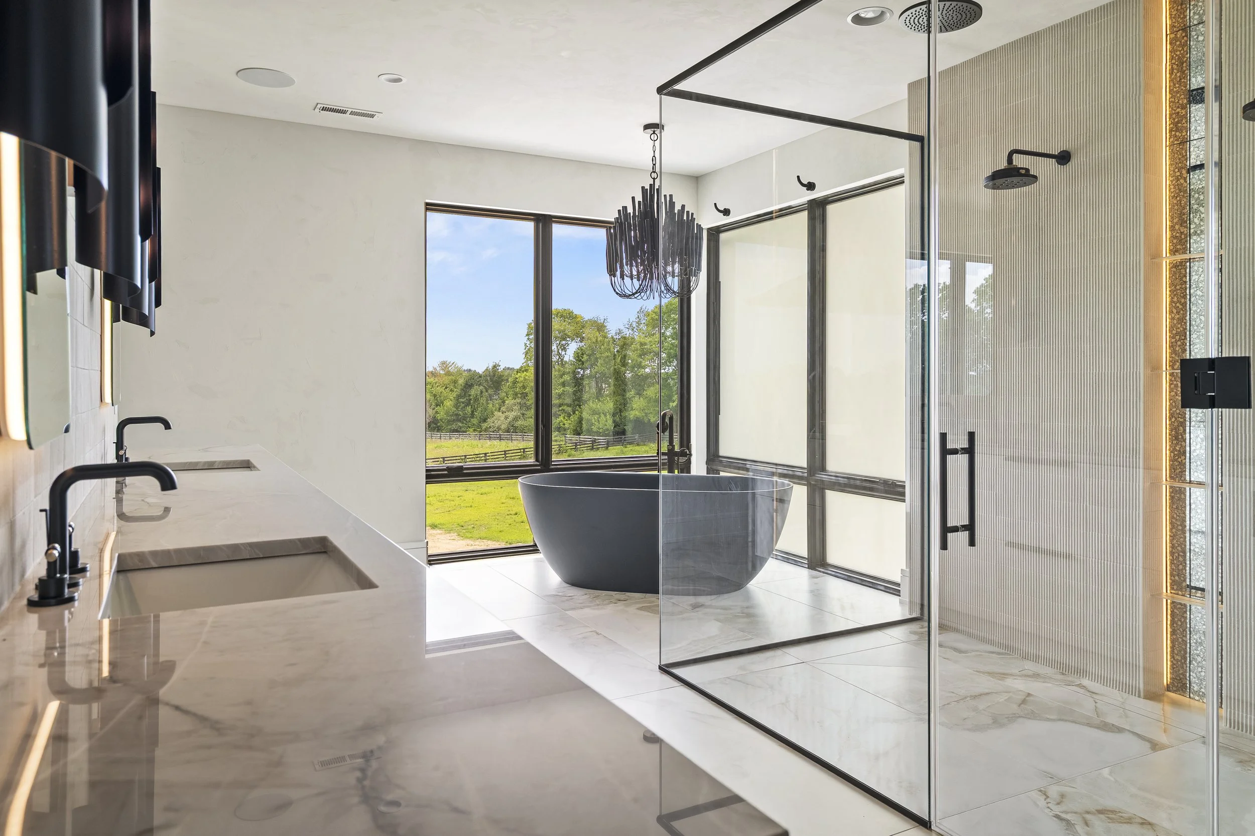 Modern bathroom with freestanding bathtub, black fixtures, glass shower enclosure, large window showcasing green landscape, chandelier, and light-colored marble floors.