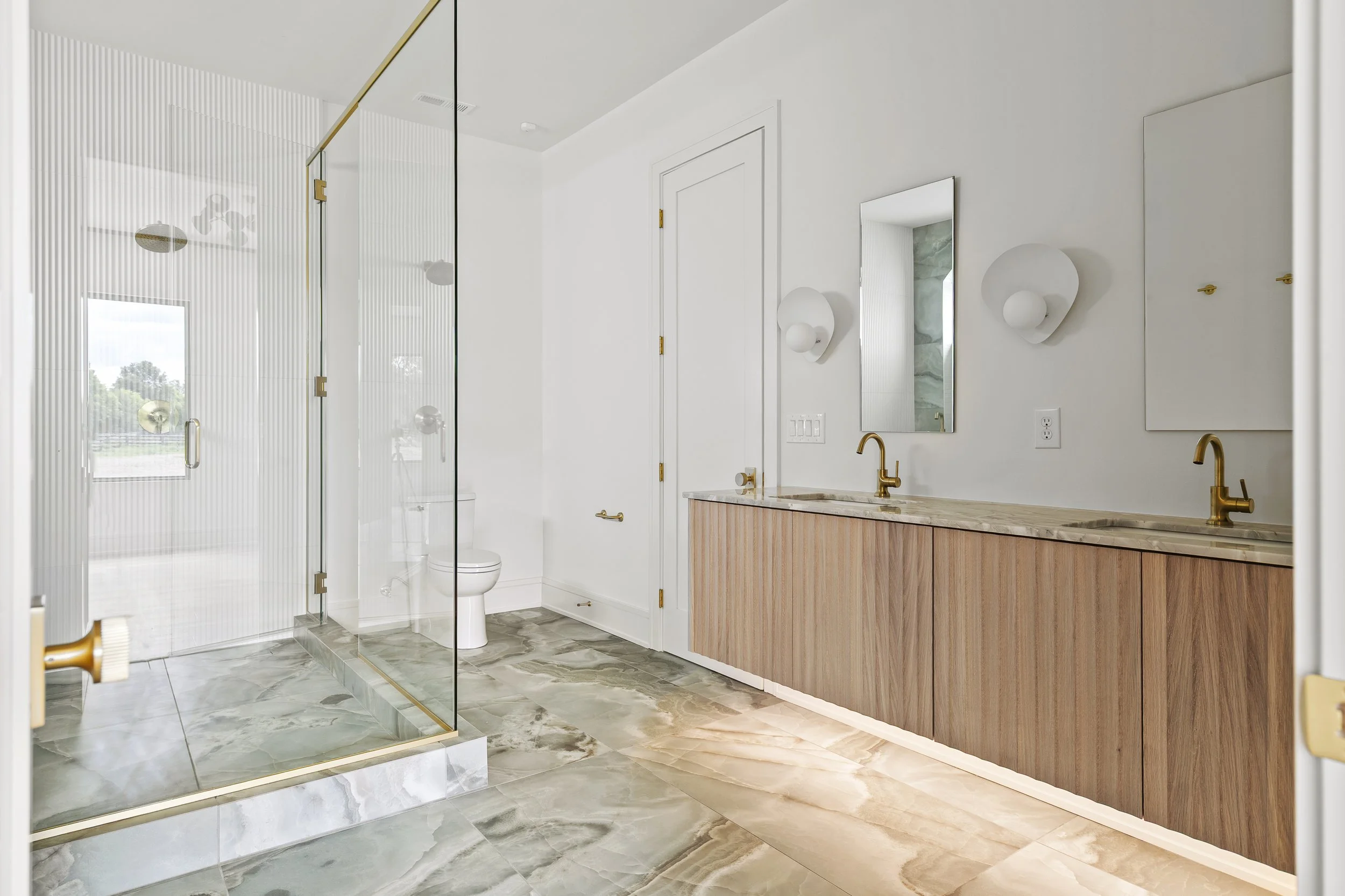 Modern bathroom with a glass shower, white walls, wooden vanity with two sinks, and gold fixtures.