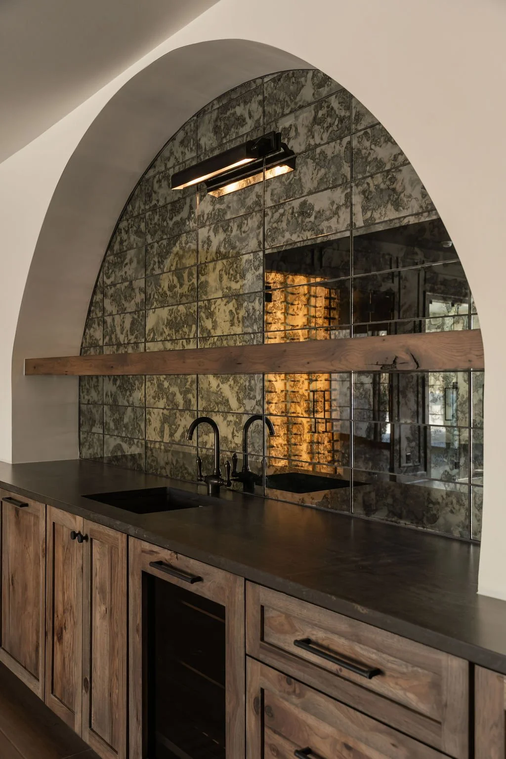 Kitchen with wooden cabinets, dark countertop, black sink, arched mirror wall feature, and industrial-style lighting.