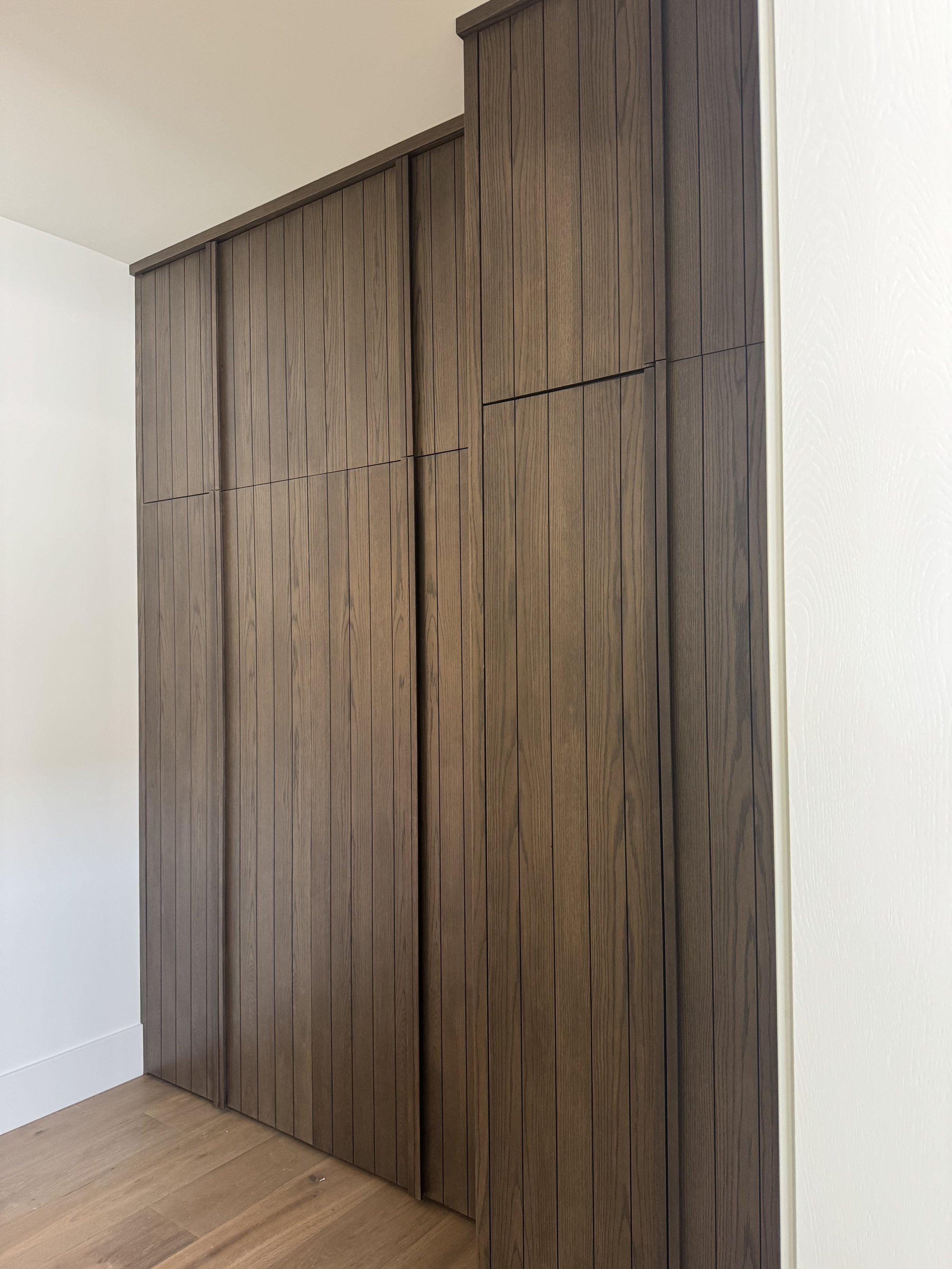 A wooden closet with vertical paneling against a white wall in a room with light wood flooring.