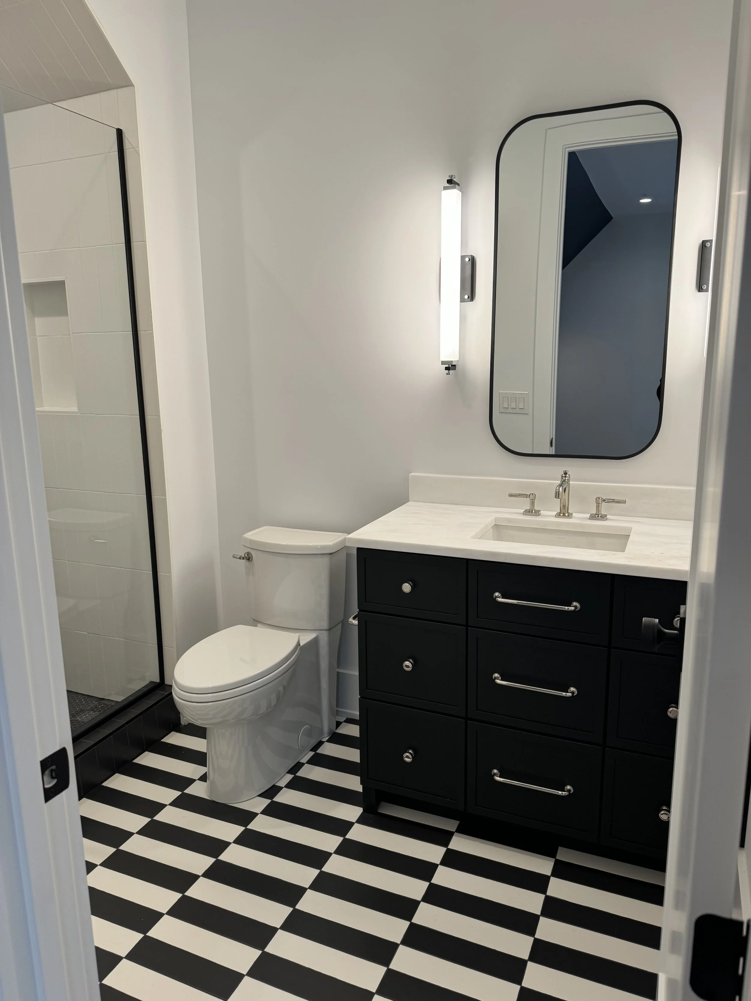 A modern bathroom with a black and white striped floor, white walls, a black vanity with silver handles, a white countertop with an integrated sink, a mirror with a black border, a wall light, a toilet, and a glass shower enclosure.