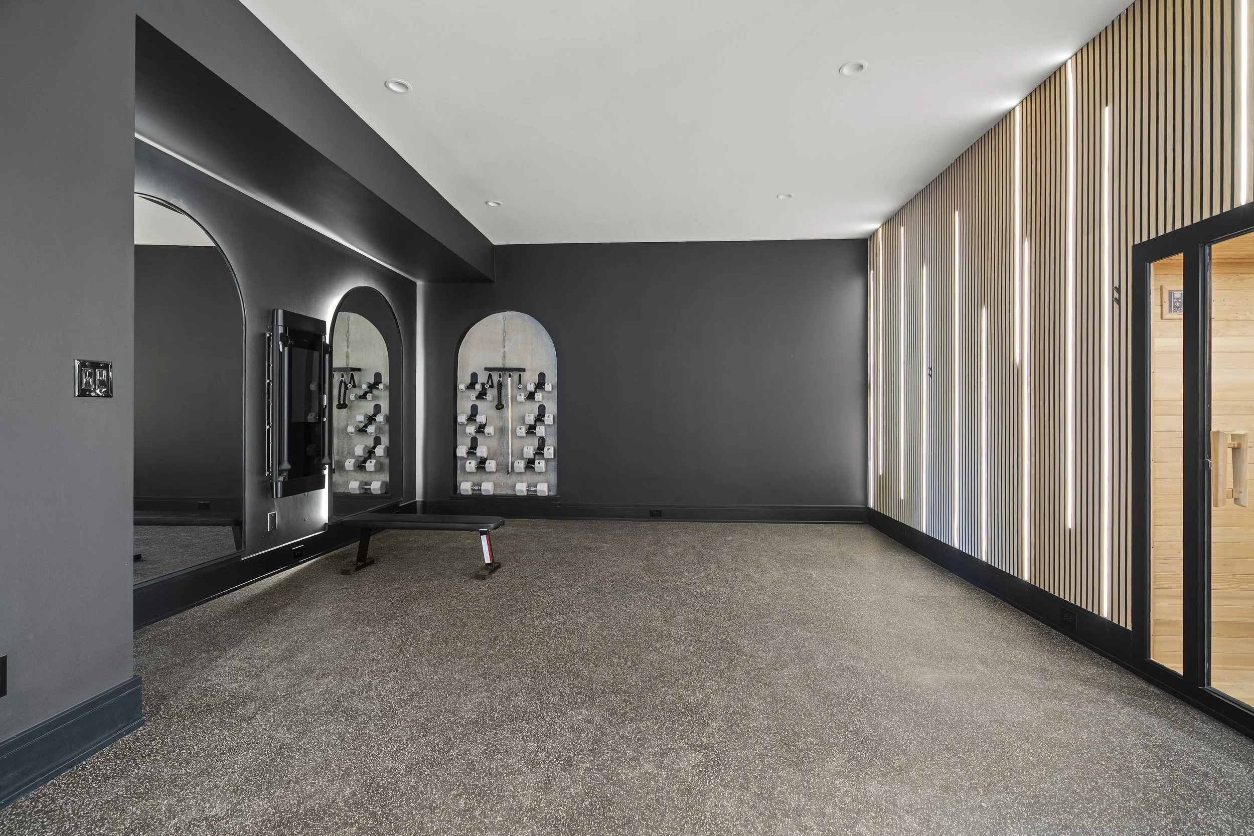 Modern interior room with black walls, mirrored workout stations, light wood paneling with vertical LED lights, gray carpeted floor, and a doorway leading to another space.