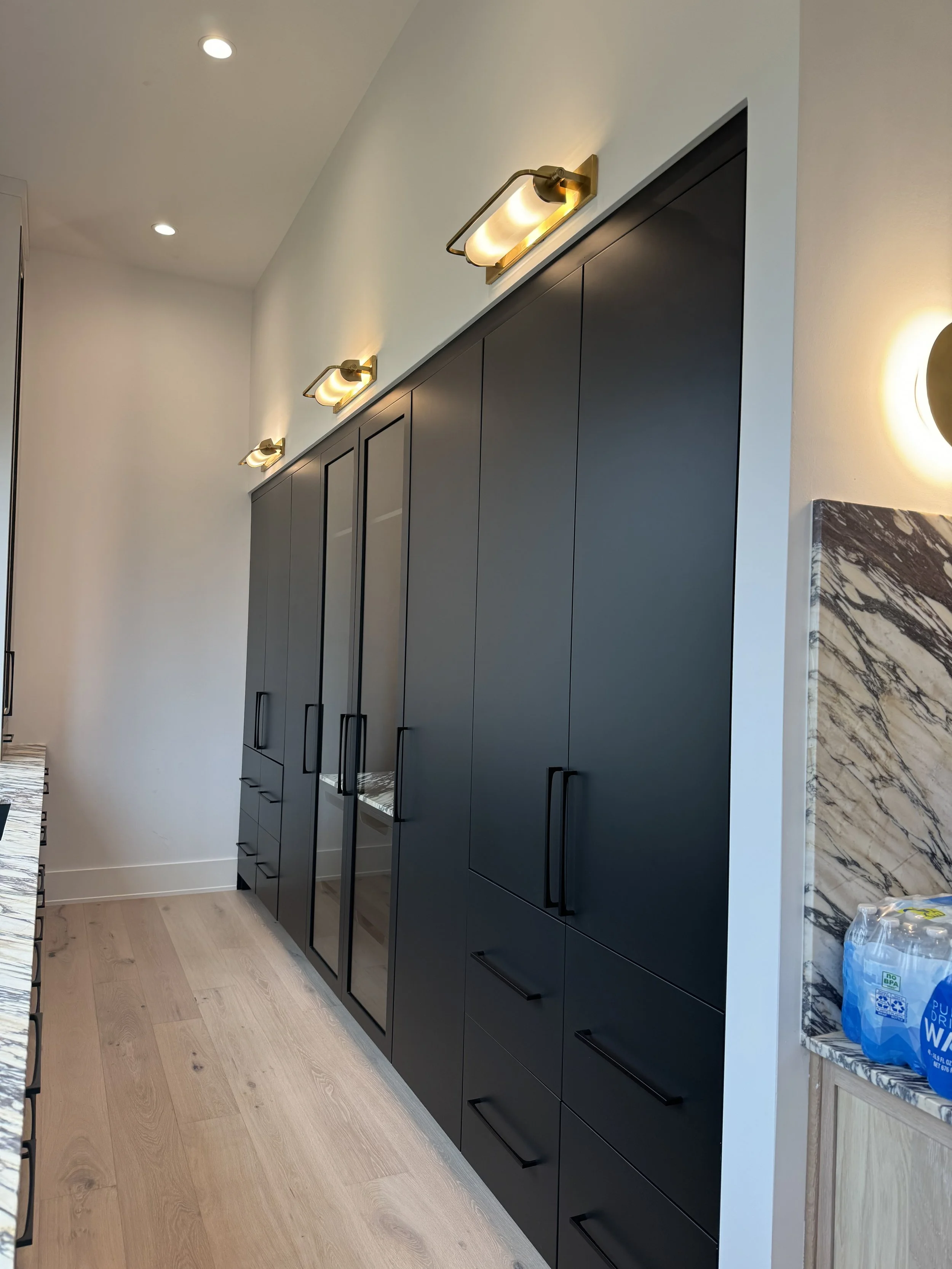 Modern kitchen with black cabinetry, light wood flooring, and marble accents. Overhead lighting fixtures and some bottled water on the counter.