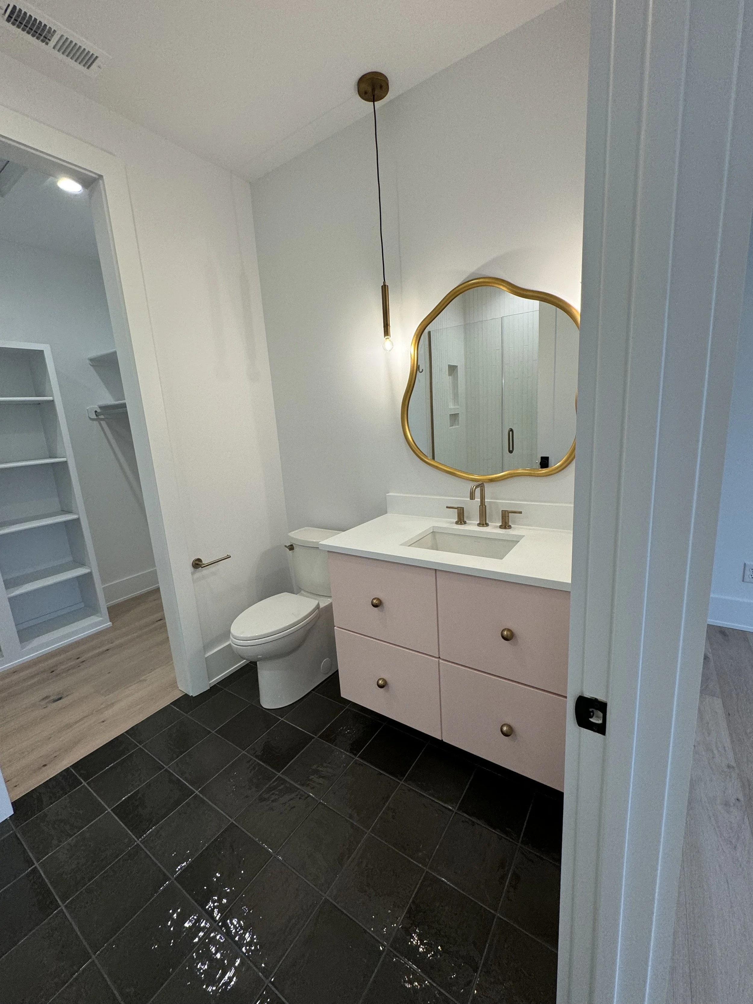 A modern bathroom with black tiled floor, white walls, a pink vanity with brass hardware, a gold-framed irregularly shaped mirror, a hanging light, and a white toilet.
