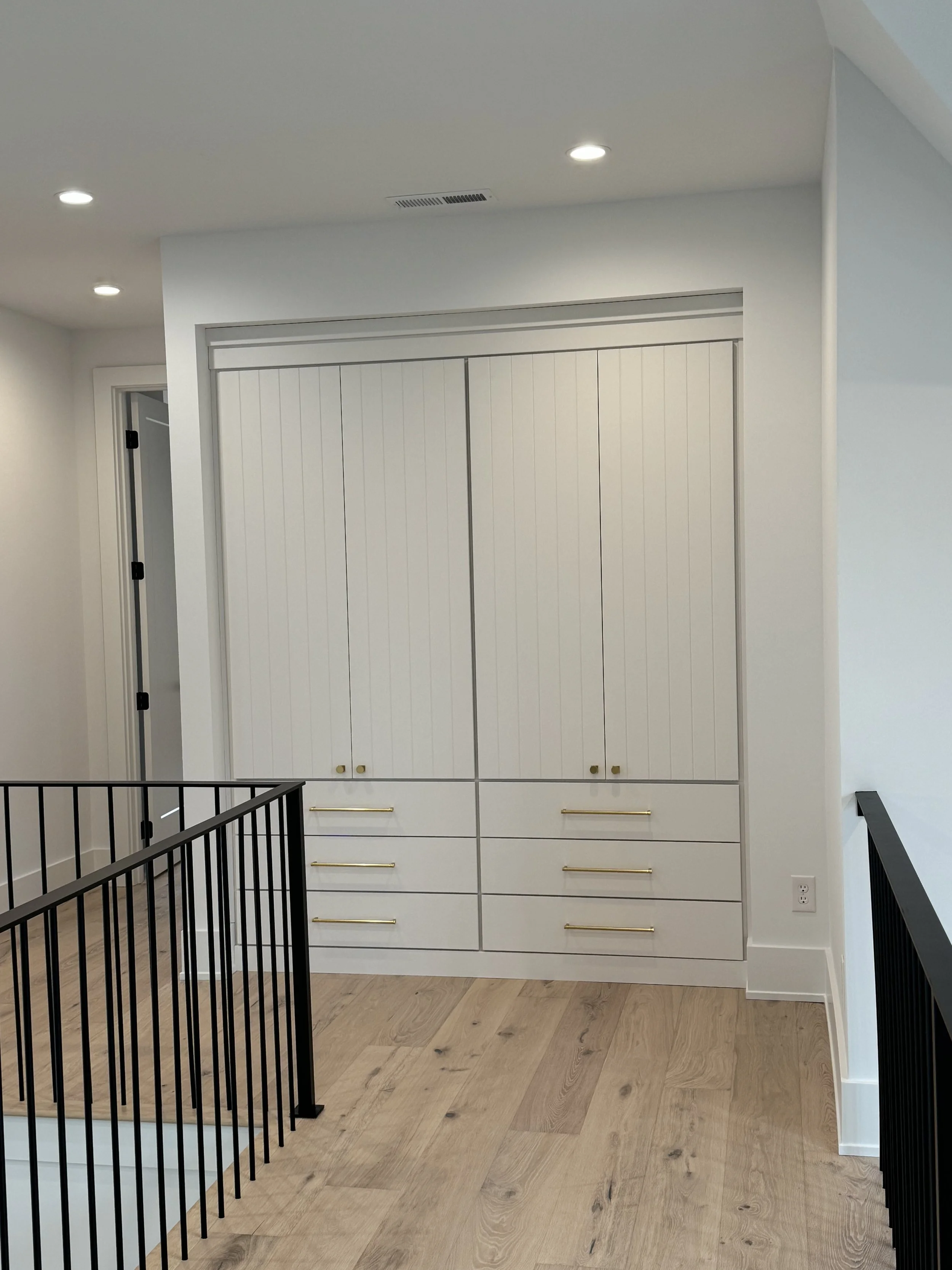 A modern white built-in closet with gold handles, located against a white wall in a room with wooden flooring. There are black metal railings in the foreground.