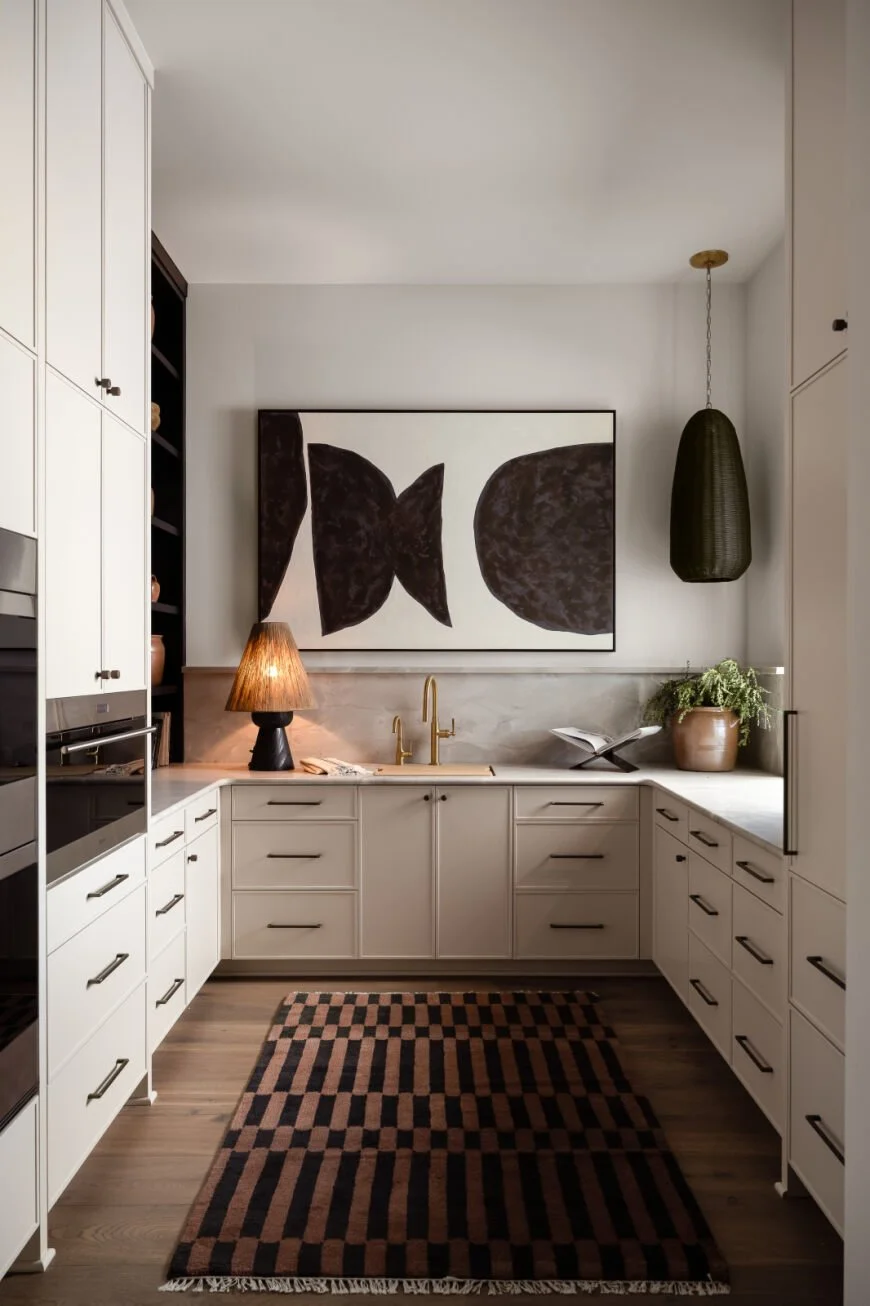 Modern kitchen with white cabinets, black open shelving, a black and beige checkered rug, gold faucet, black pendant light, decorative lamp, potted plant, and abstract black and white artwork.