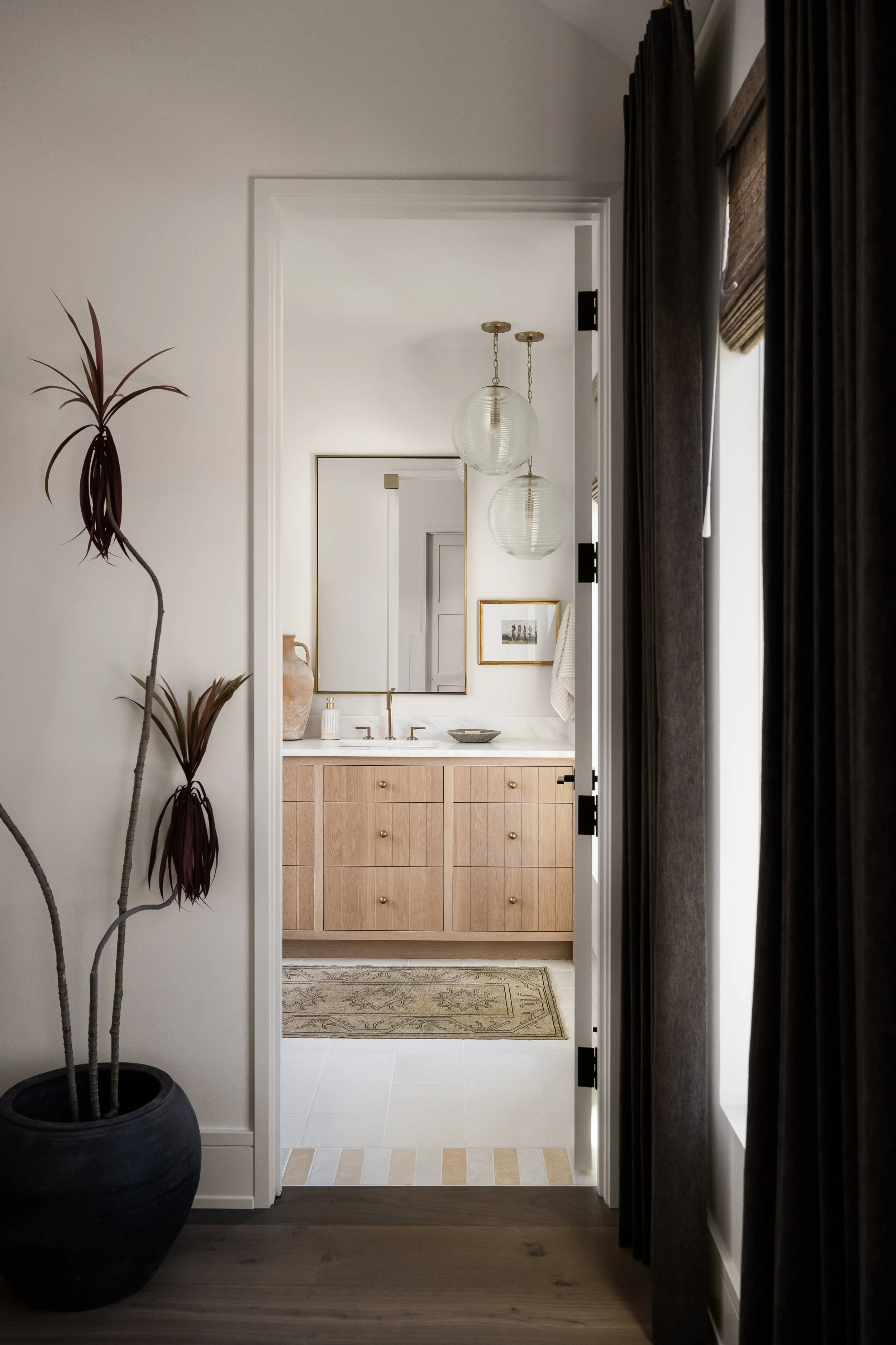 A view through doorway into a bathroom with a wooden vanity, a mirror, and hanging globe pendant lights.
