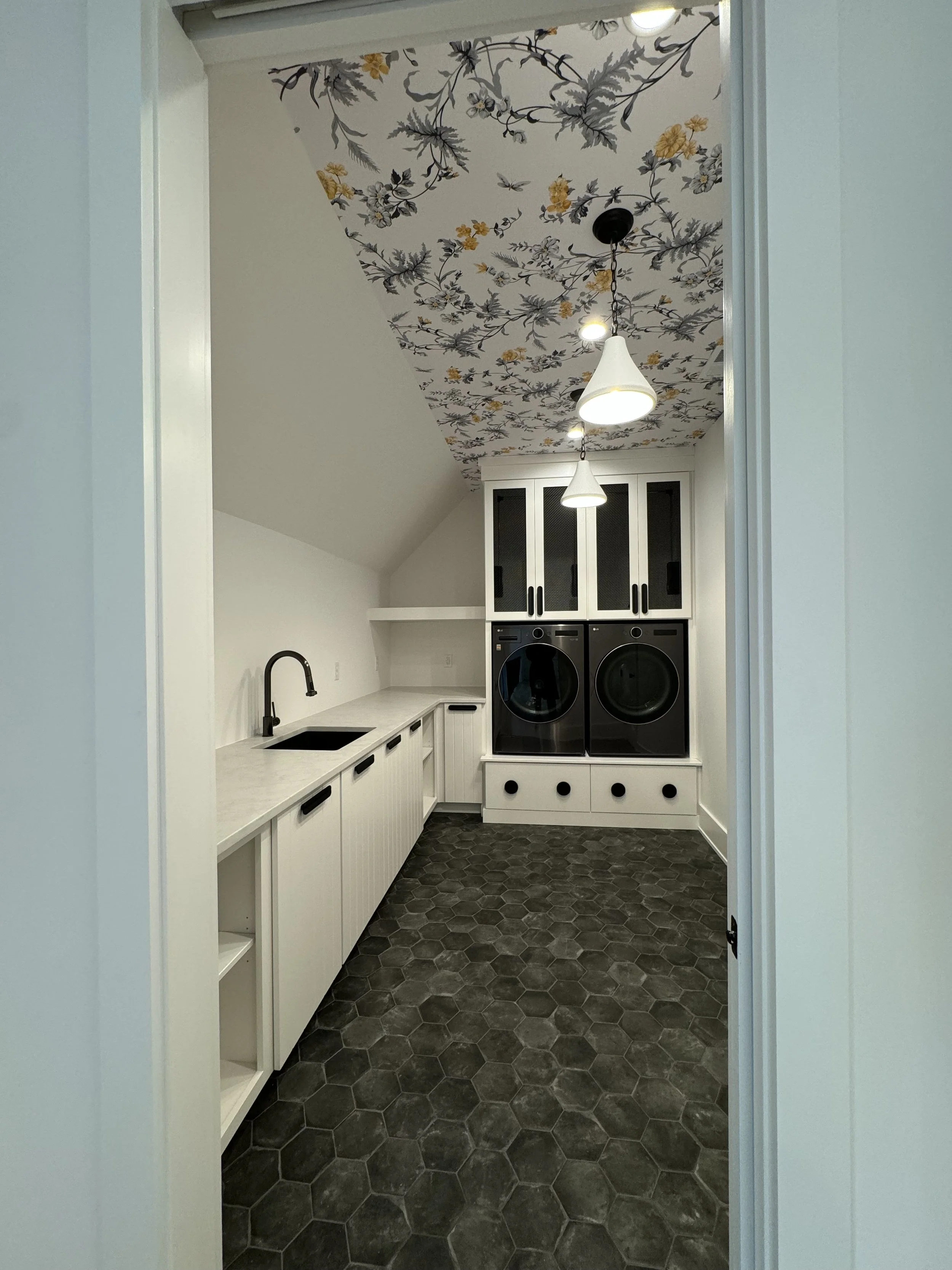 A laundry room viewed through a doorway, featuring white cabinets with black handles, a black washer and dryer, a floral patterned ceiling with gray and yellow flowers, and dark hexagonal tile flooring.