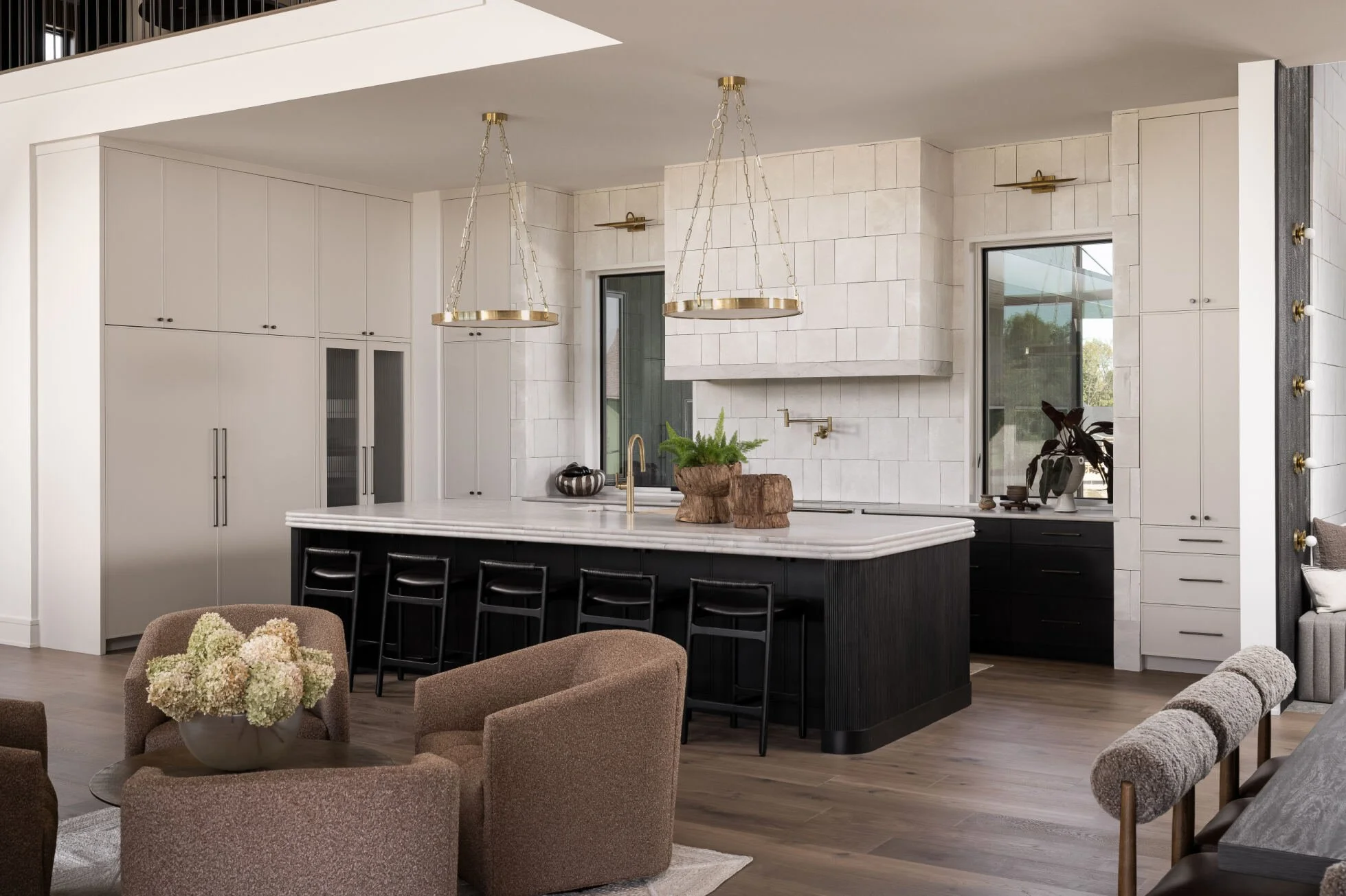 Modern kitchen with white cabinets, black island, gold pendant lights, and a marble countertop. There are windows and various decorative items, including a plant and flowers.