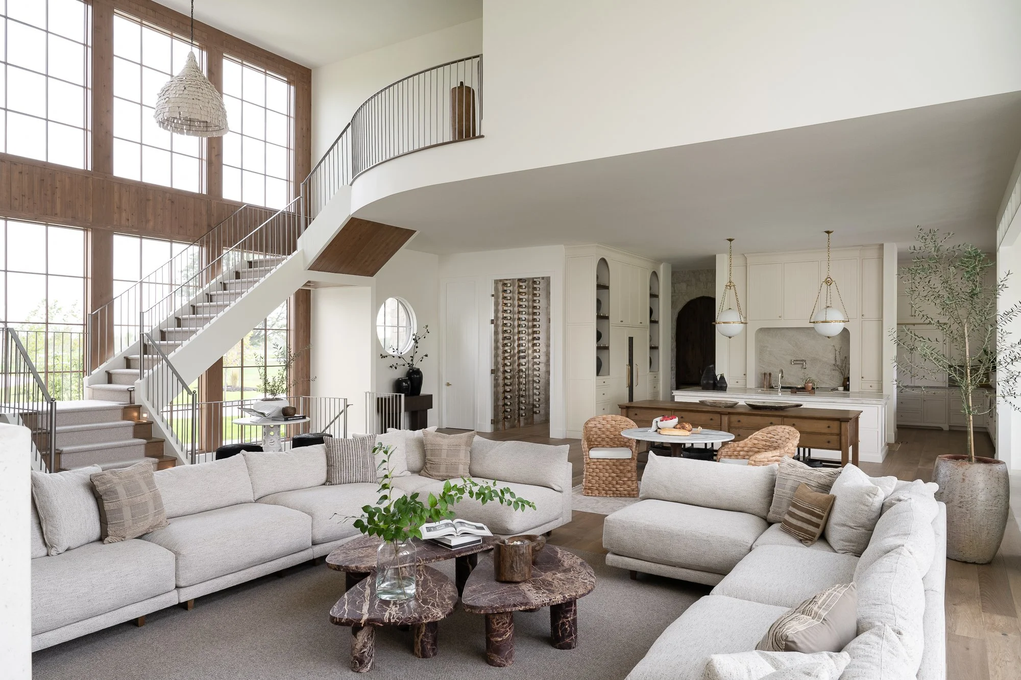 Interior of a spacious modern living room with large windows, a white couch, coffee tables, and a kitchen area in the background.