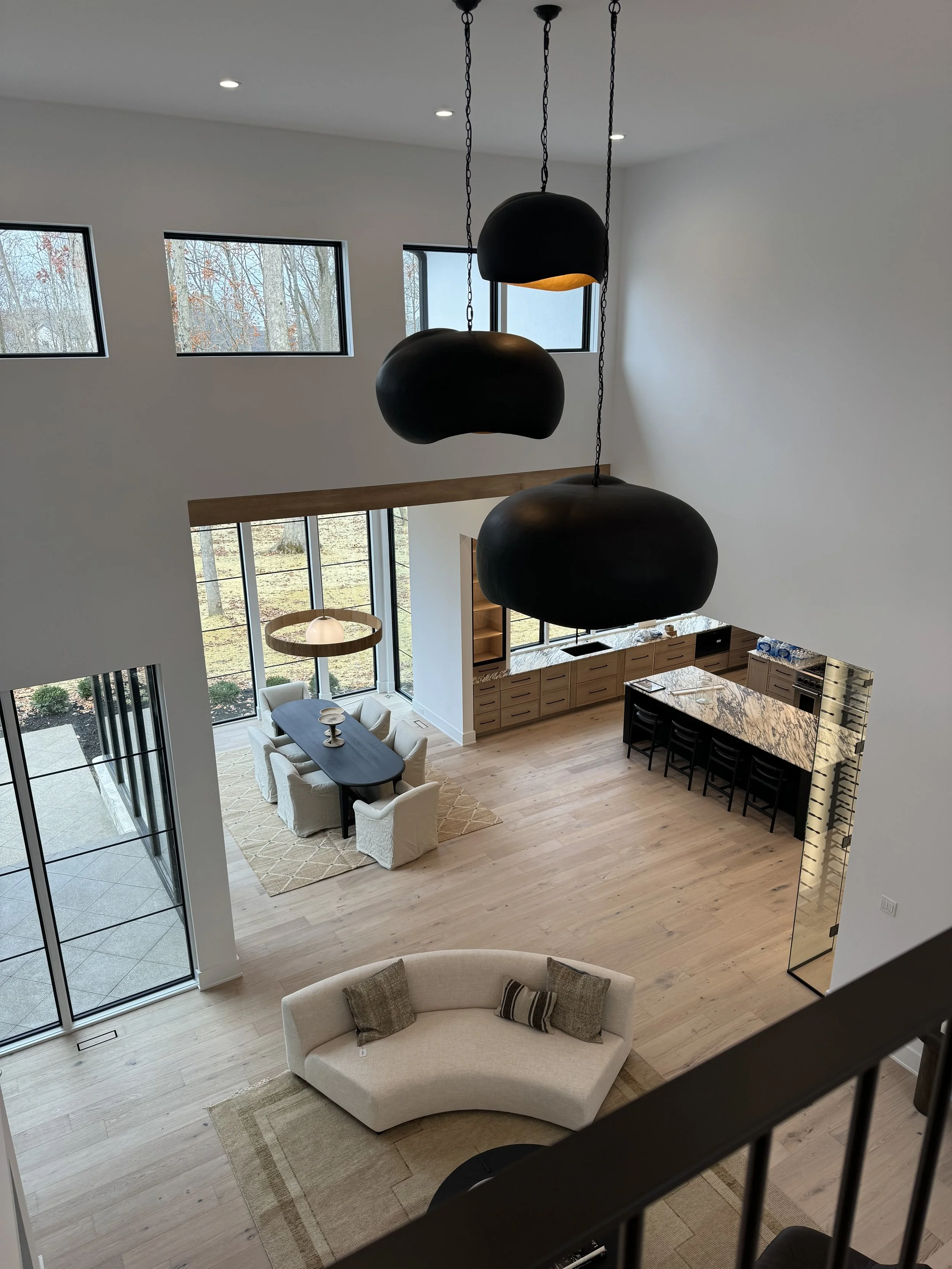Living room with high ceiling, three black pendant lights, large windows showing trees outside, a dining table with white chairs, a beige sofa with throw pillows, and kitchen area with wooden cabinets and a marble countertop.