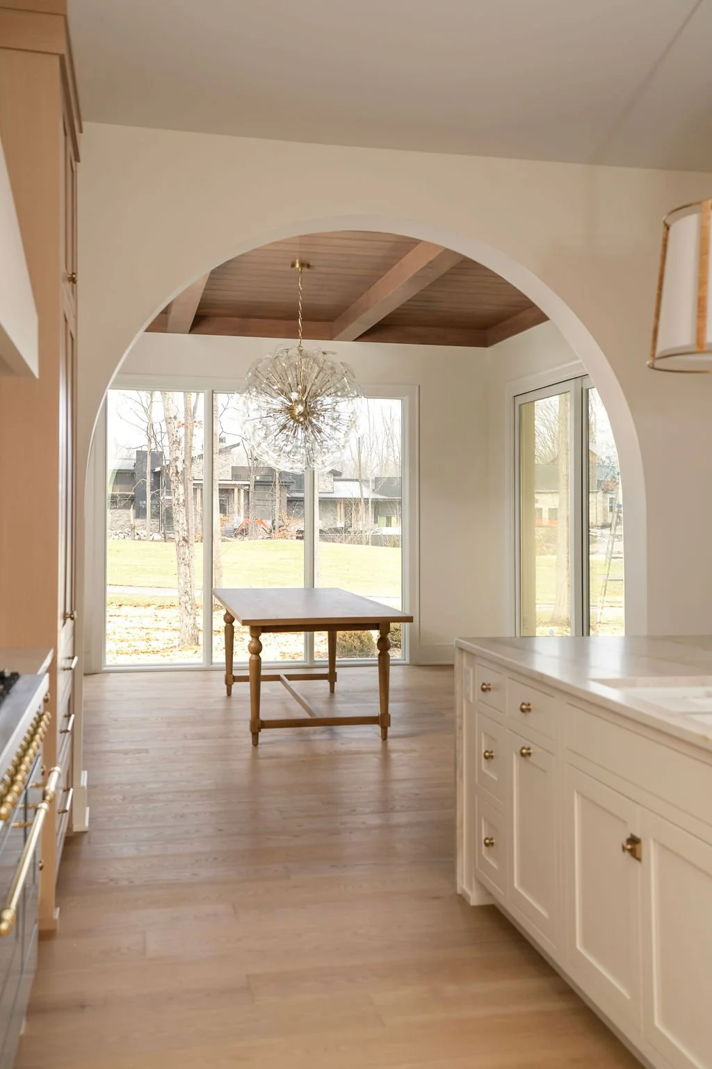 A bright kitchen with large windows, a wooden table, a chandelier, and white cabinetry.