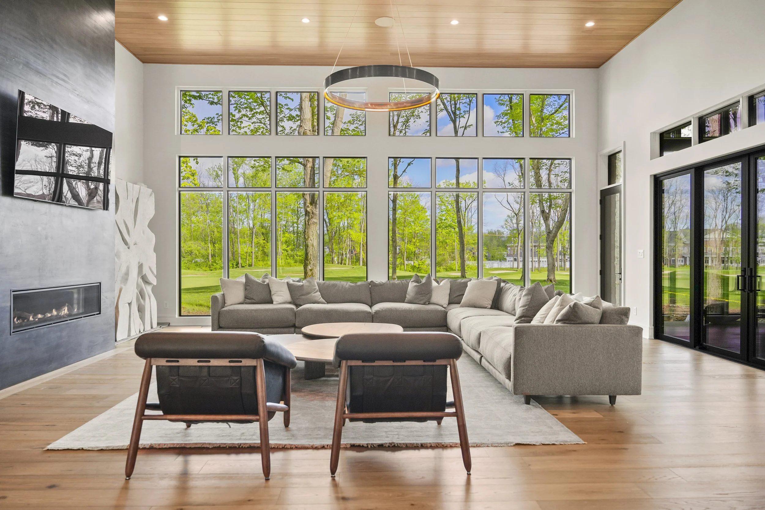 A spacious living room with large windows showing green trees outside. There is a gray sectional sofa with beige pillows, a black chair with a wooden frame, a light-colored rug, and a modern circular hanging light fixture on a wooden ceiling.
