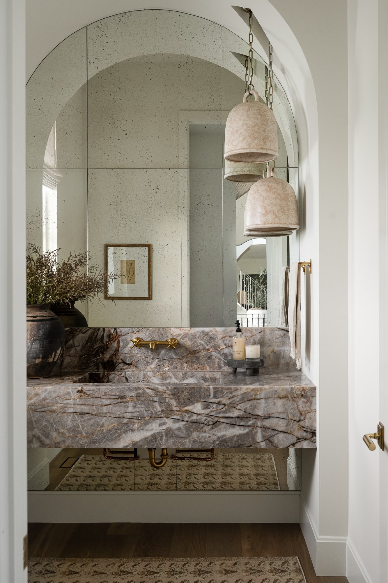 A stylish bathroom with a marble vanity countertop, a large mirror, pendant lights, and a vase with greenery.