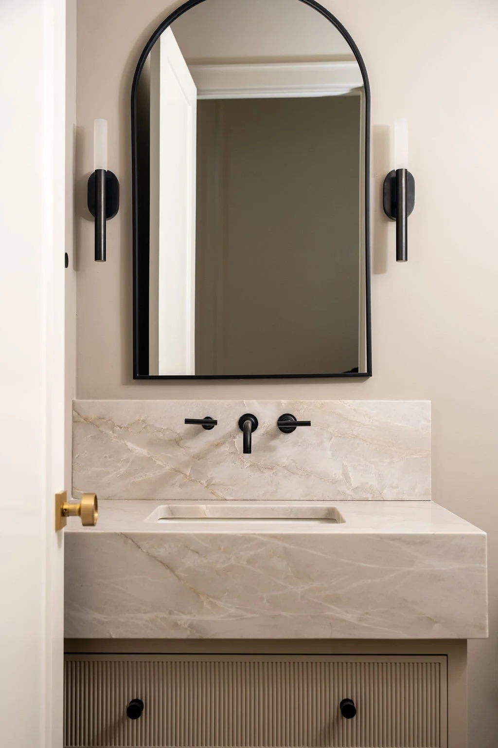 Bathroom vanity with a marble countertop, black faucet, matching black wall-mounted light fixtures on each side, a large arched mirror, and a beige wall.