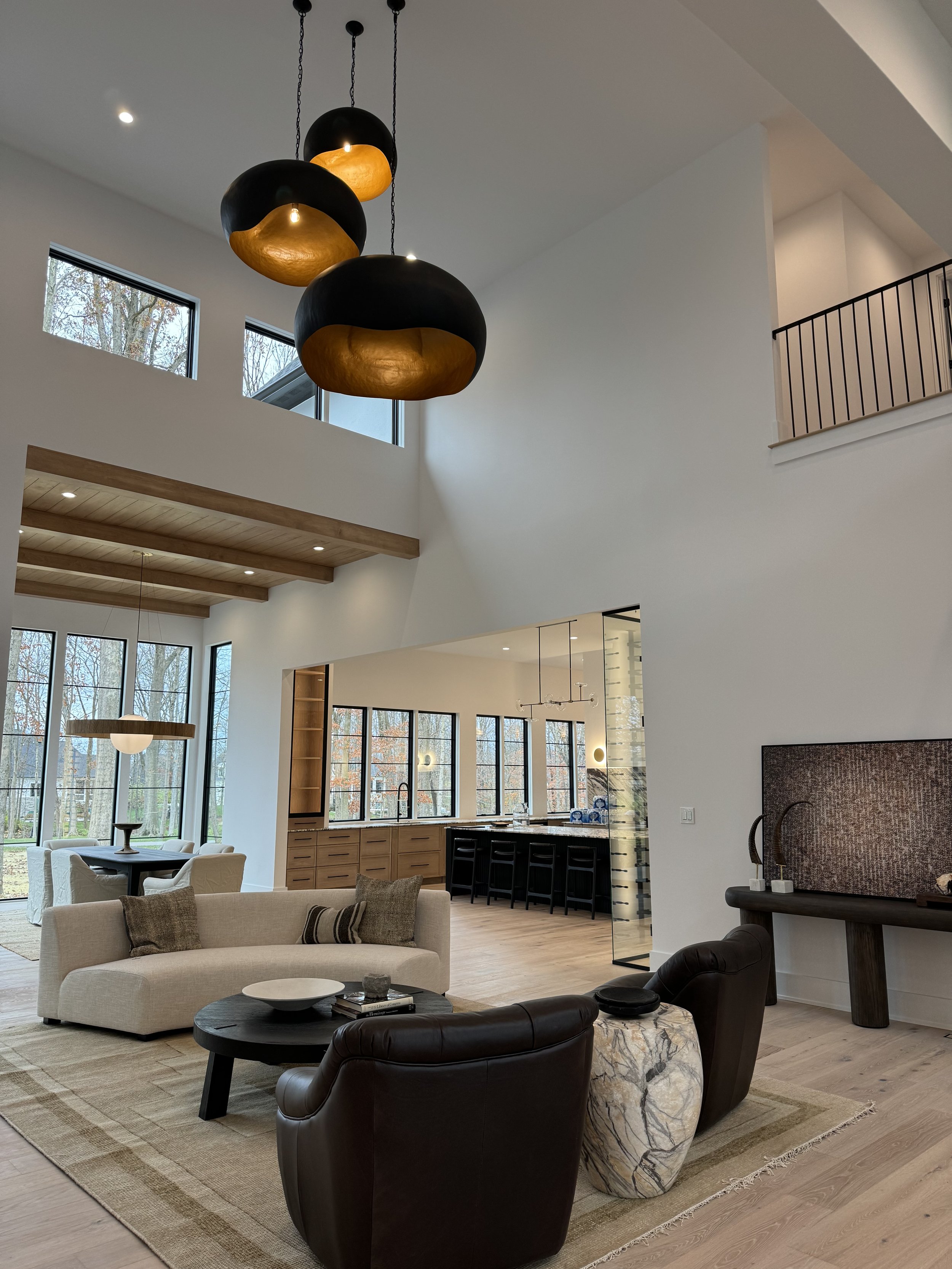 Modern open-concept living room with high ceilings, large windows, and contemporary furniture. Features include a beige sofa with pillows, a black leather armchair, a marble side table, a dark coffee table, and a patterned area rug. The background sh