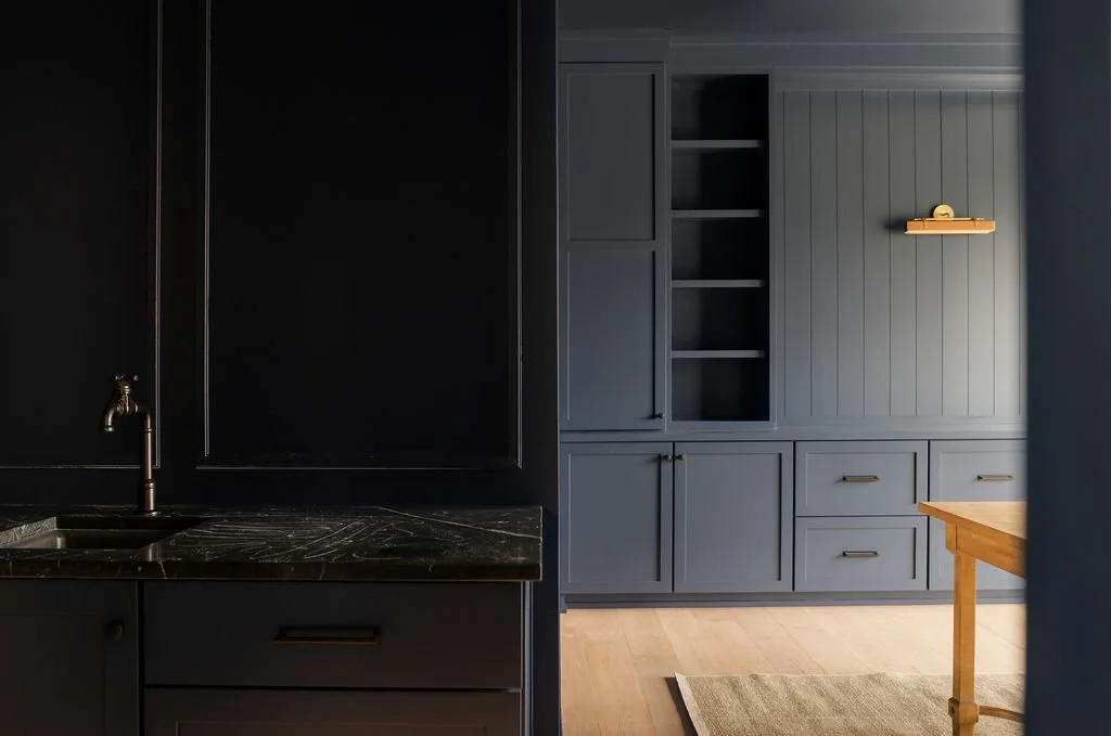 A modern kitchen with dark blue cabinetry, a black countertop with marble pattern, brass hardware, and a built-in shelving unit in blue. Part of a wooden table and light hardwood floor are visible.