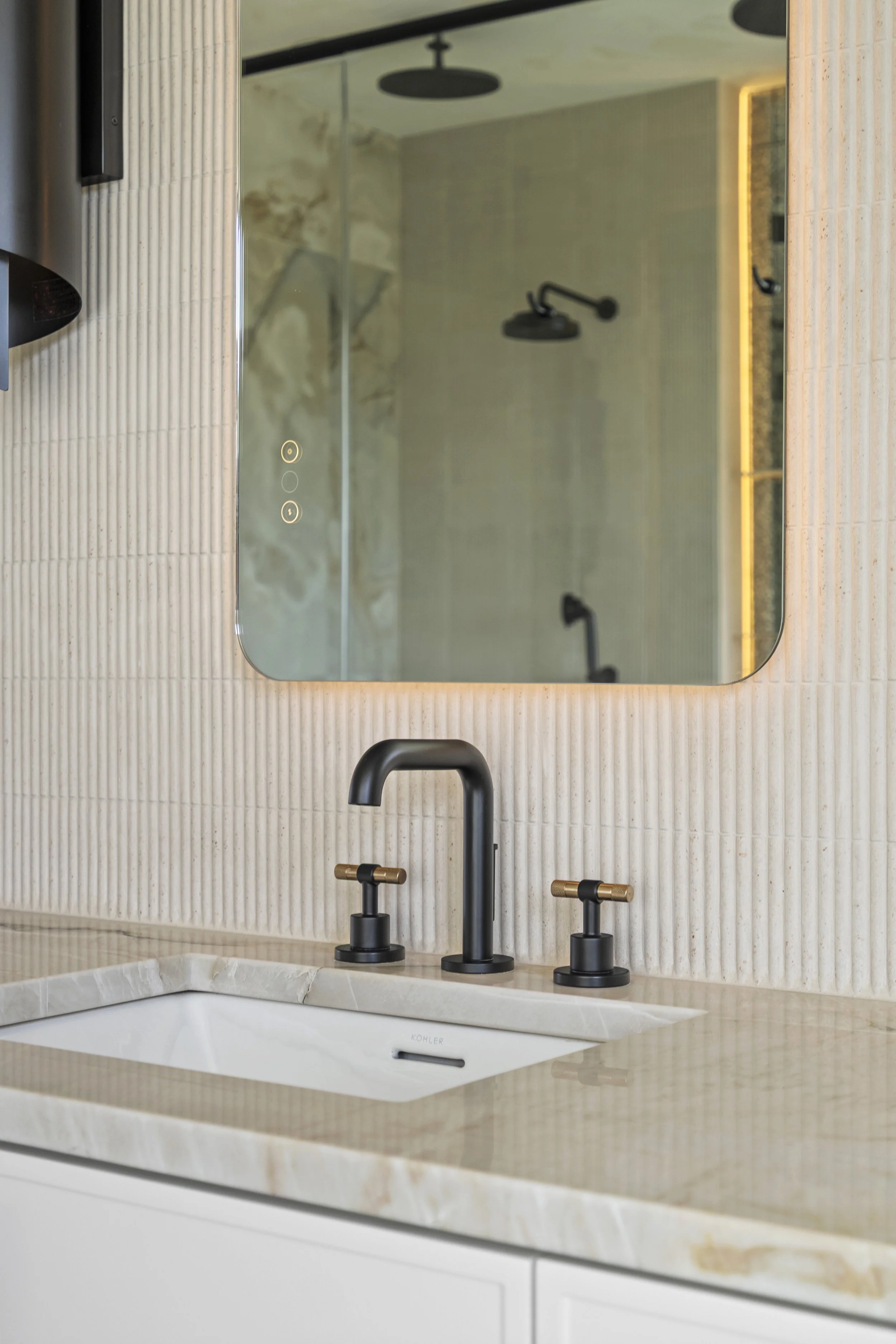 Close-up of bathroom countertop with a white sink, black faucet, and black handles, a large mirror, and part of a shower with black fixtures in the reflection.
