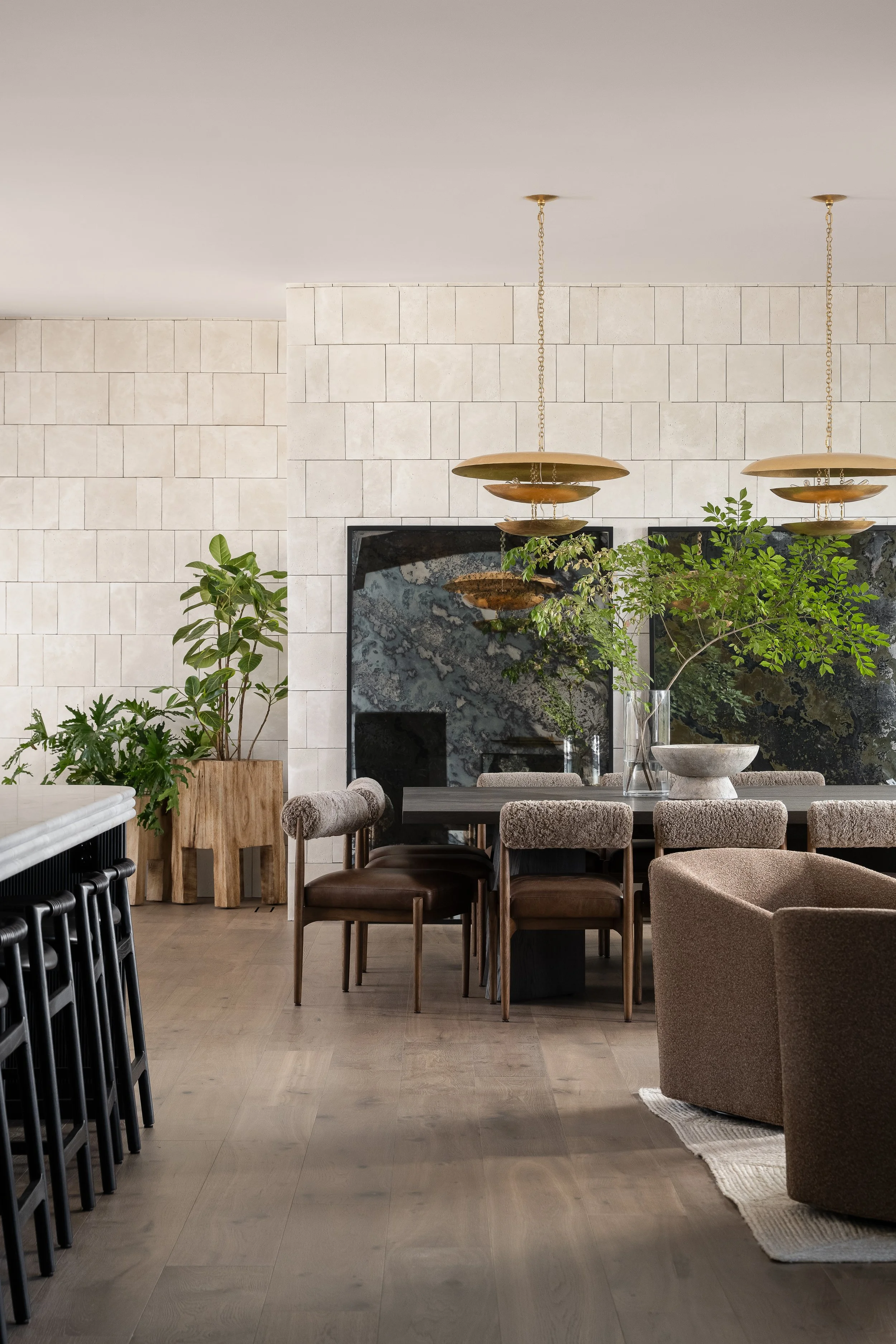 A modern dining space with a light wood floor, a half wall made of light-colored stone, and a white ceiling. There are several green potted plants, including a large one in a wooden planter. Hanging above the table are two modern brass pendant lights